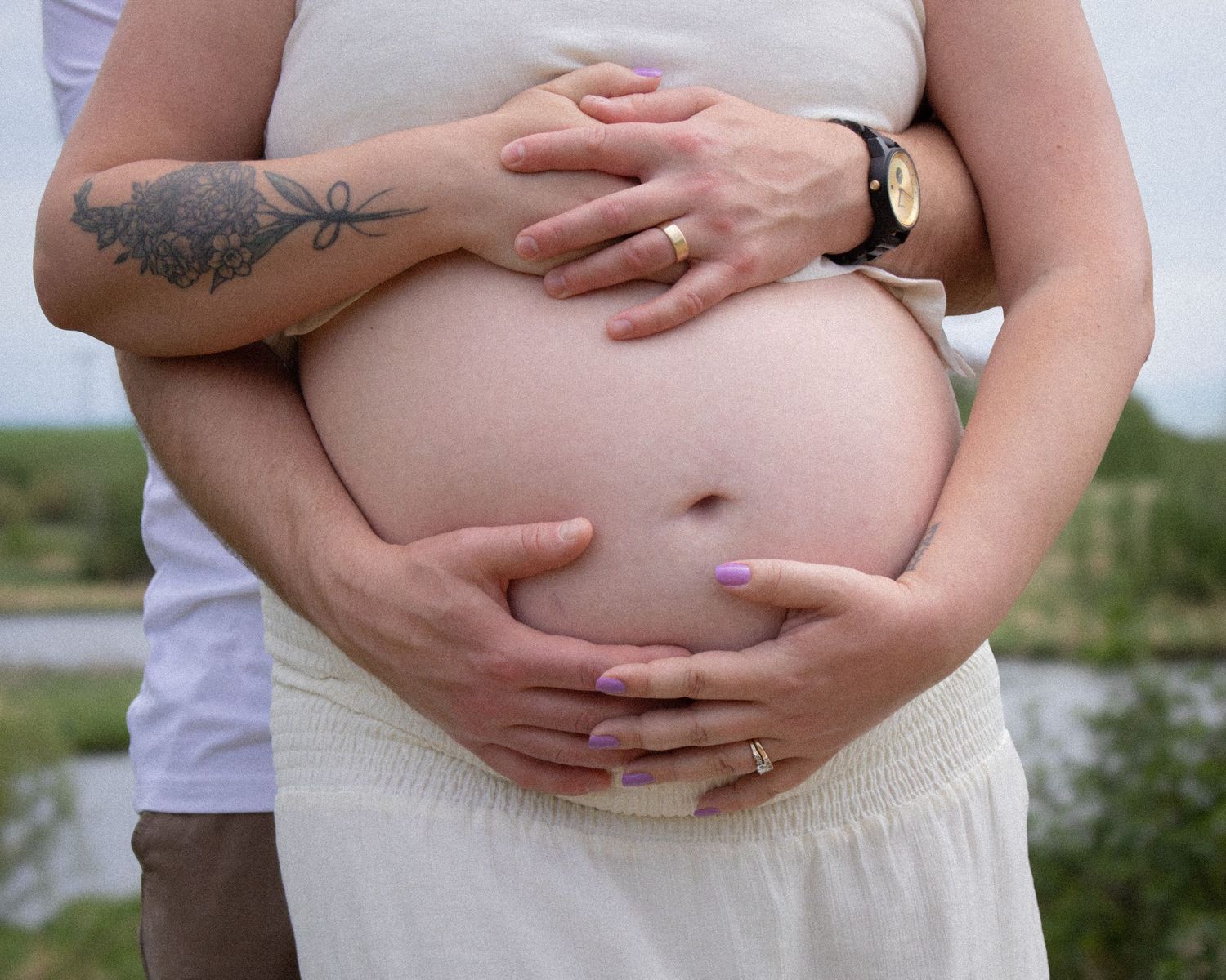 Pregnant woman in white dress with partner's hands cradling baby bump outdoors near water.