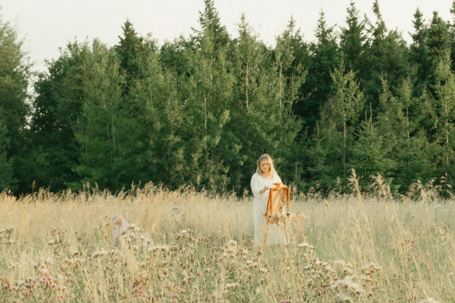 Blonde woman in white top and brown skirt standing in a golden field surrounded by tall green trees.