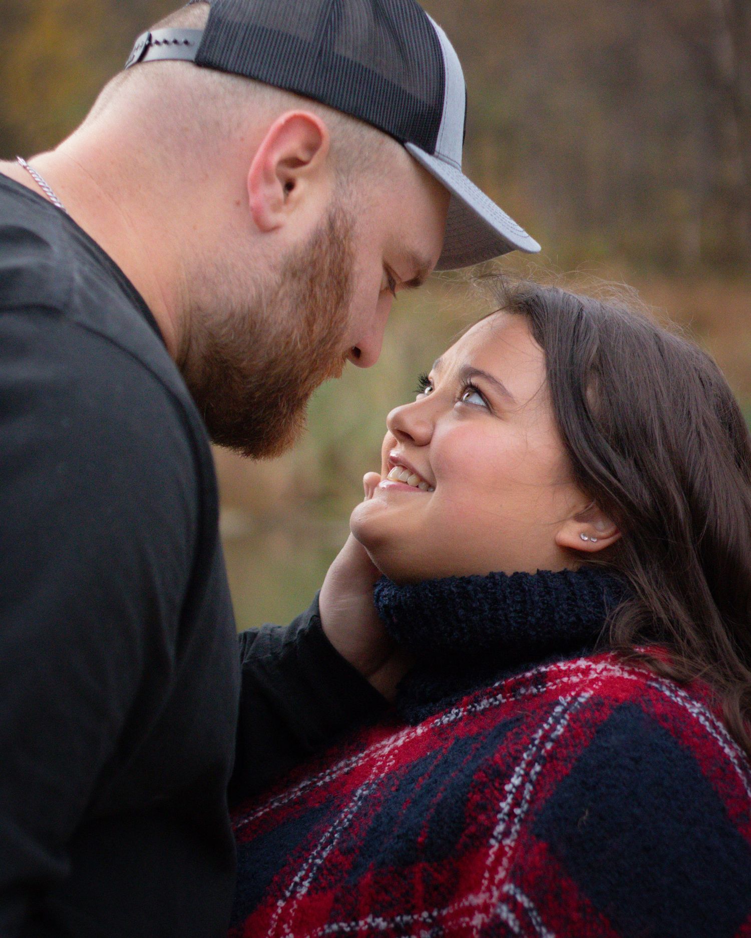 Romantic couple sharing a tender moment outdoors in autumn, woman smiling up at bearded man in snapback cap.