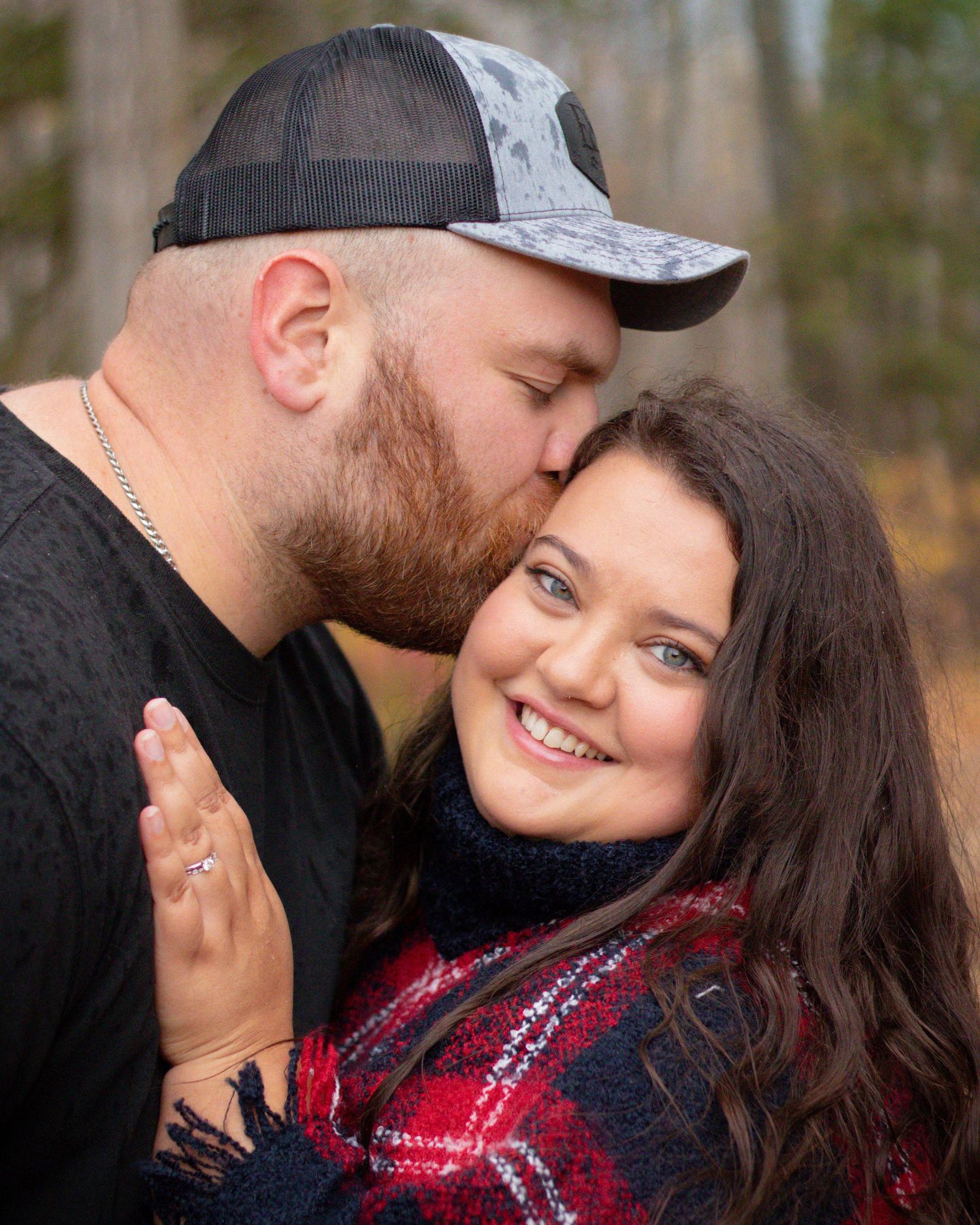 Bearded man kissing smiling woman showing engagement ring in autumn forest setting.