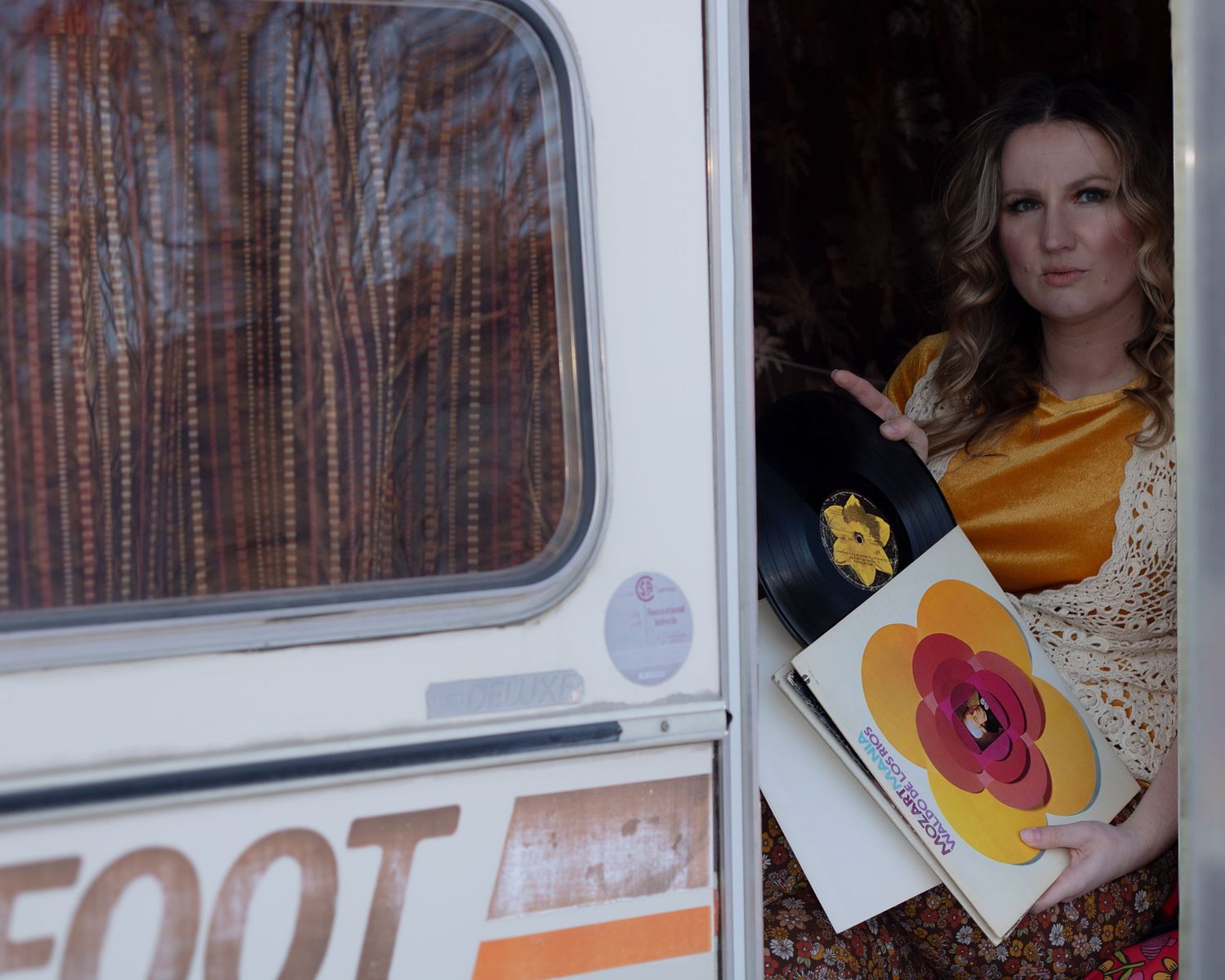 Woman in gold and lace dress displaying vinyl record album from vintage Bigfoot camper doorway.