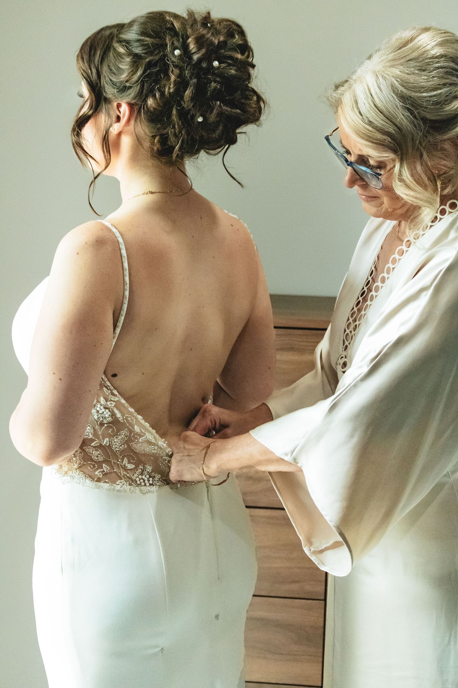Mother helping bride button the back of her elegant lace wedding dress before the ceremony.