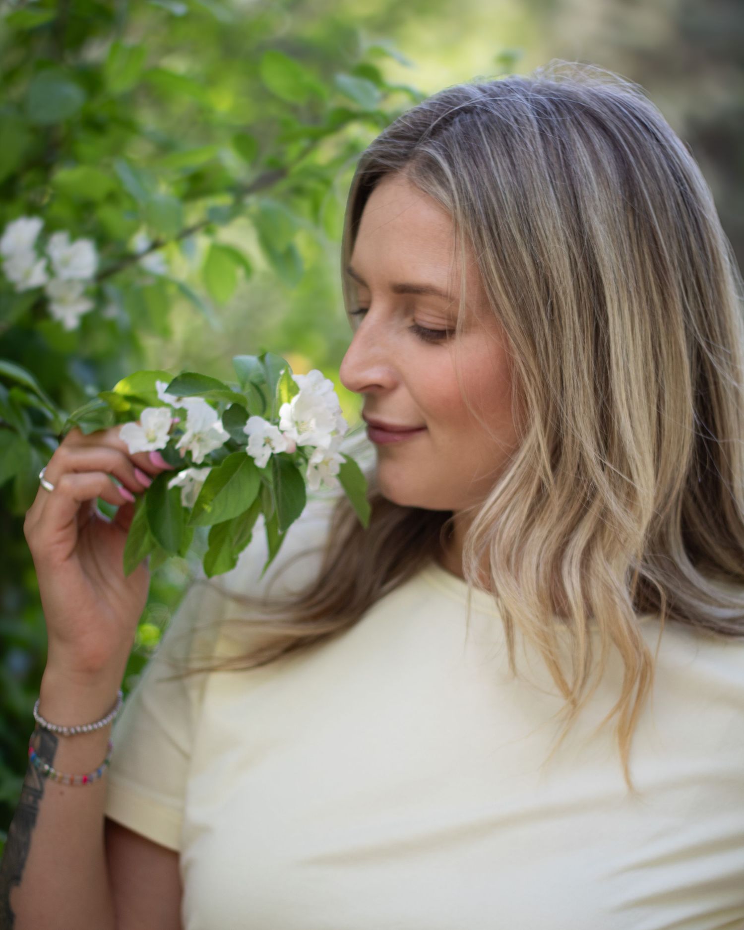 Woman with blonde hair smelling white blossoms on a tree branch outdoors in a lush green garden.