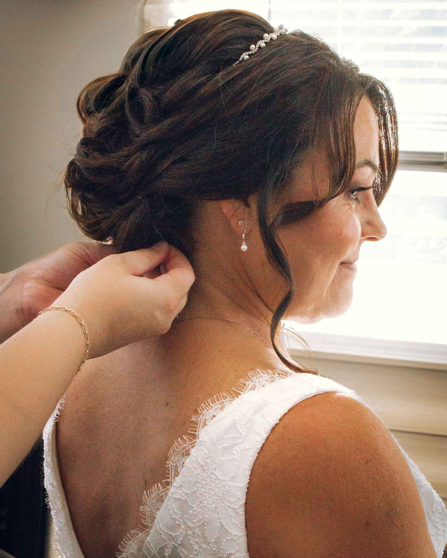 Bride smiling as someone helps her put on earrings, wearing a white lace dress and delicate hair accessories.