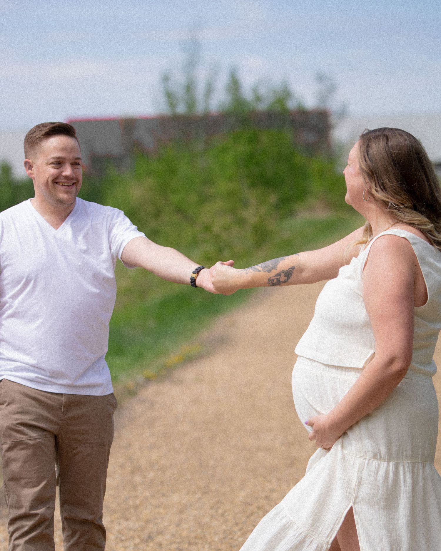 Couple holding hands on a path outdoors, pregnant woman in white dress, man in white shirt and khaki pants, sunny day.