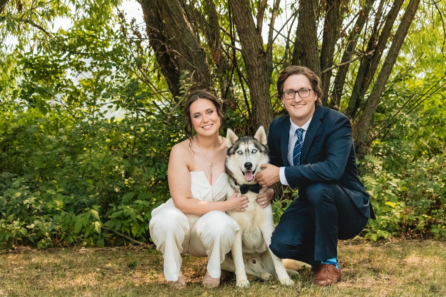 Smiling bride and groom crouching with their Siberian Husky dog outdoors on their wedding day.
