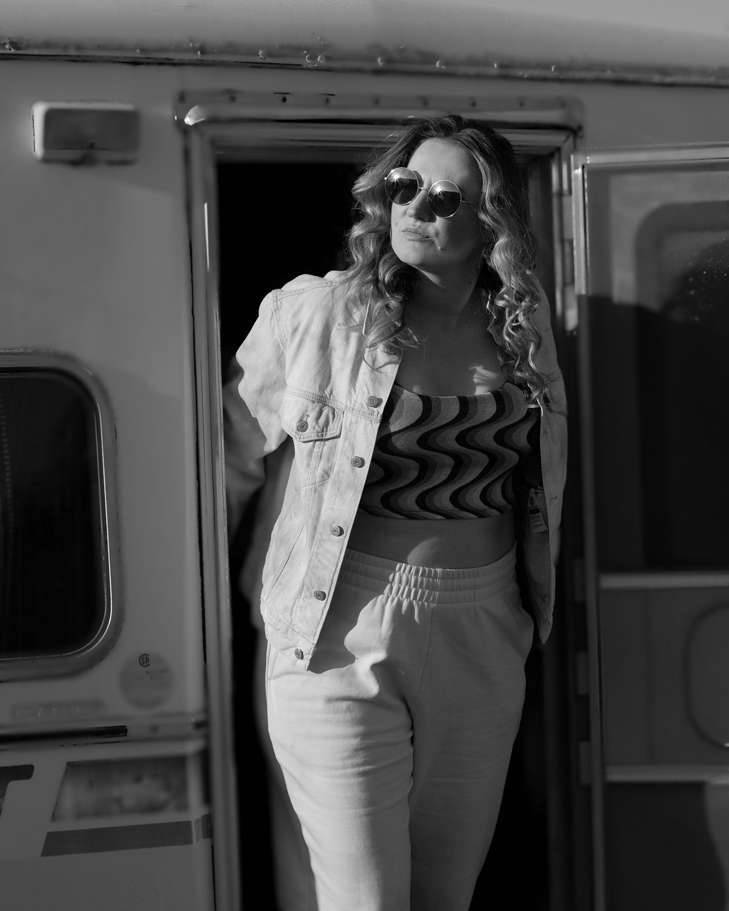 Black and white photo of woman in sunglasses and denim jacket leaning against a vintage camper door.