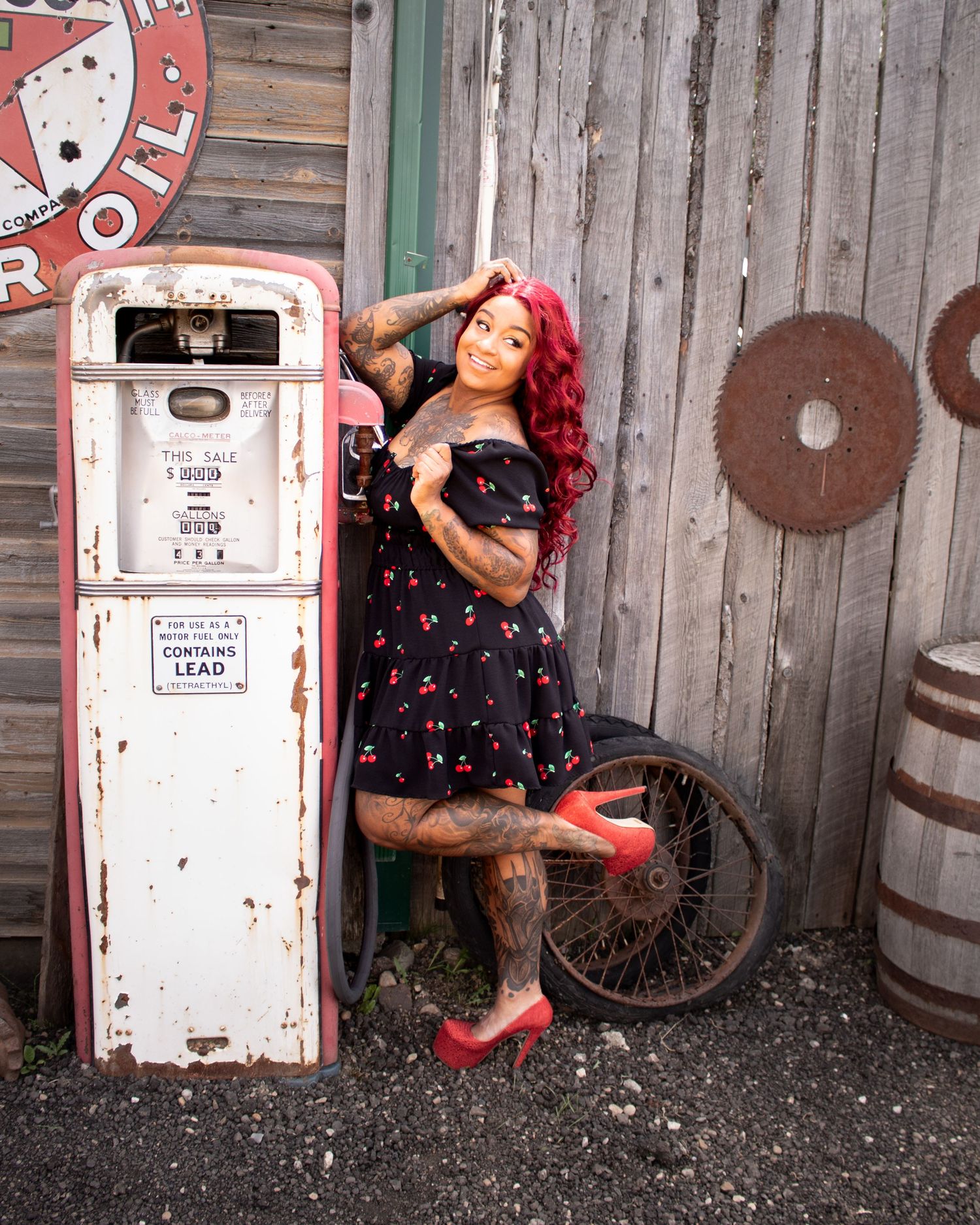 Tattooed woman with red hair in a cherry dress leaning against a vintage gas pump.