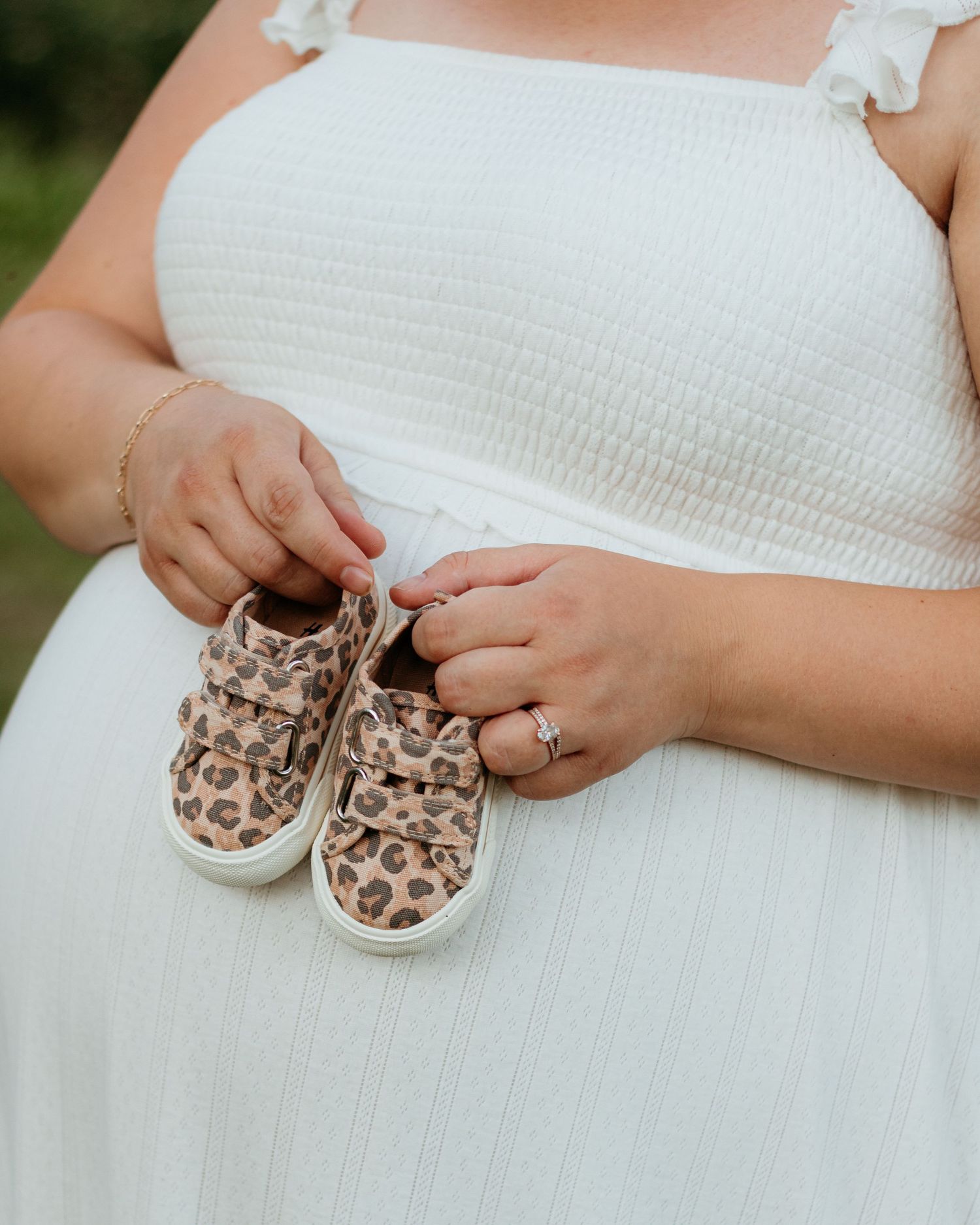 Pregnant woman in white dress holding tiny leopard print baby sneakers against her bump.