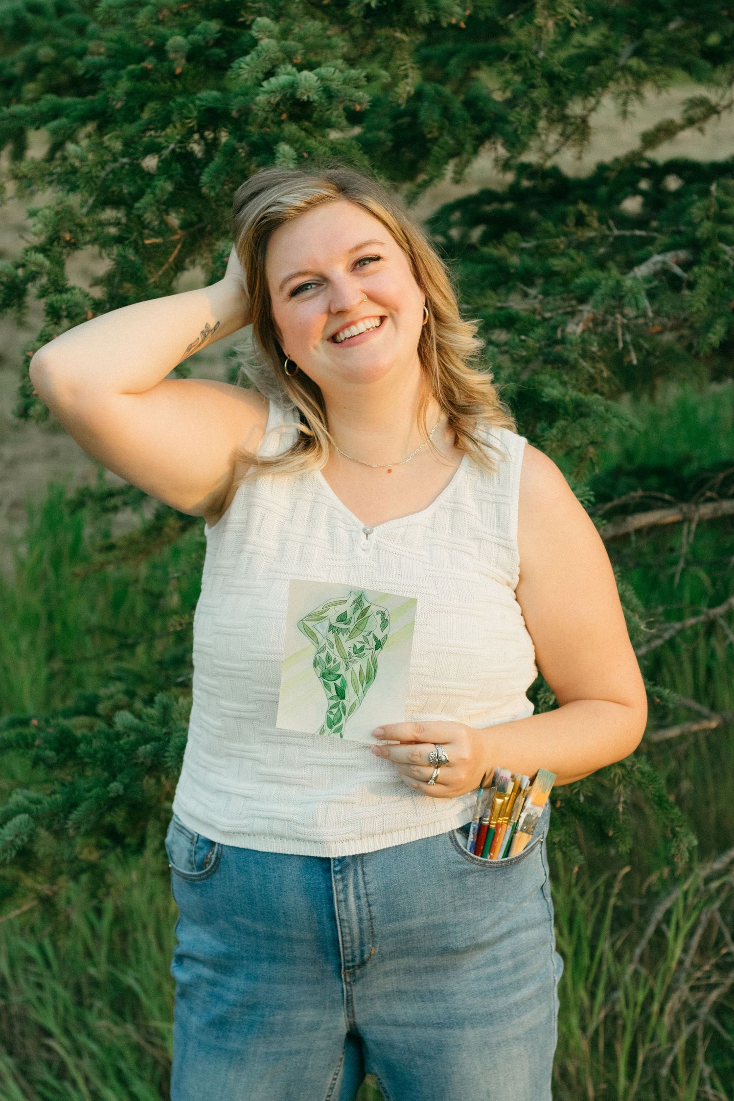 Smiling blonde woman in white tank top and jeans holding artwork and paintbrushes outdoors near green trees.