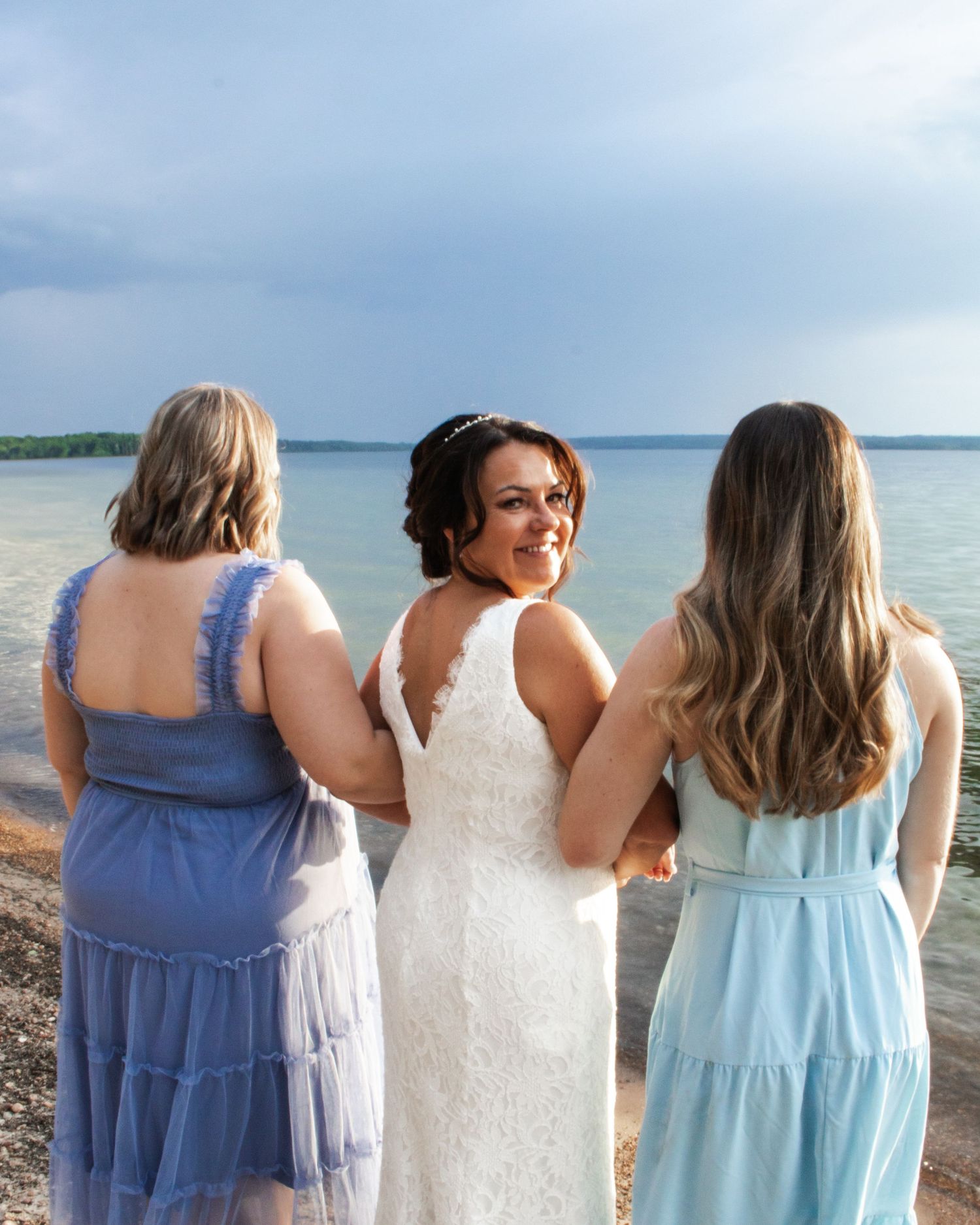 Bride in white lace dress smiles back at camera while standing with two bridesmaids in blue by a lake.