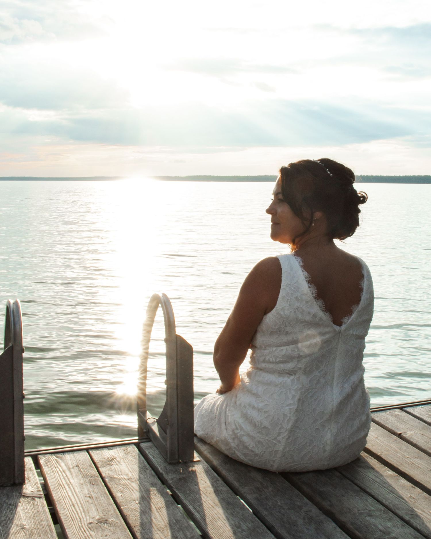 Bride in white lace dress sits on wooden dock gazing at a serene sunset over calm lake waters.