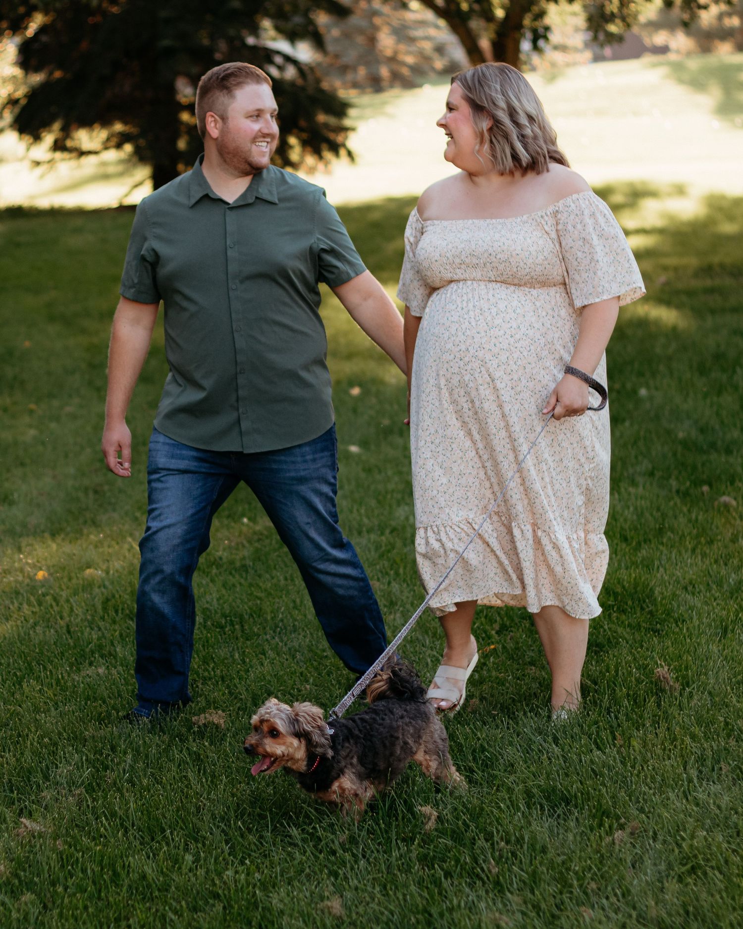 Couple holding hands walking their small dog on a leash in a green park during a maternity photo session.