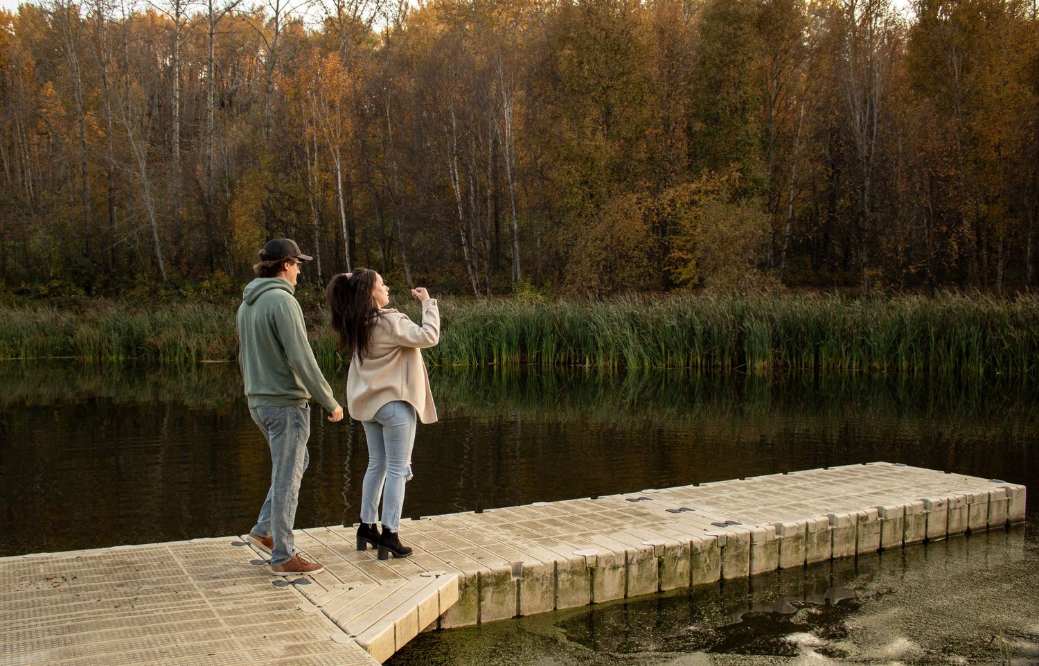 Couple holding hands on a wooden dock overlooking a calm autumn lake surrounded by fall foliage.