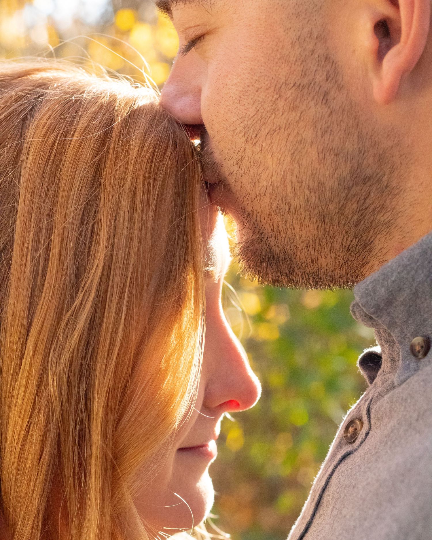 Man with beard gently kissing redhead woman on forehead in warm golden autumn sunlight.