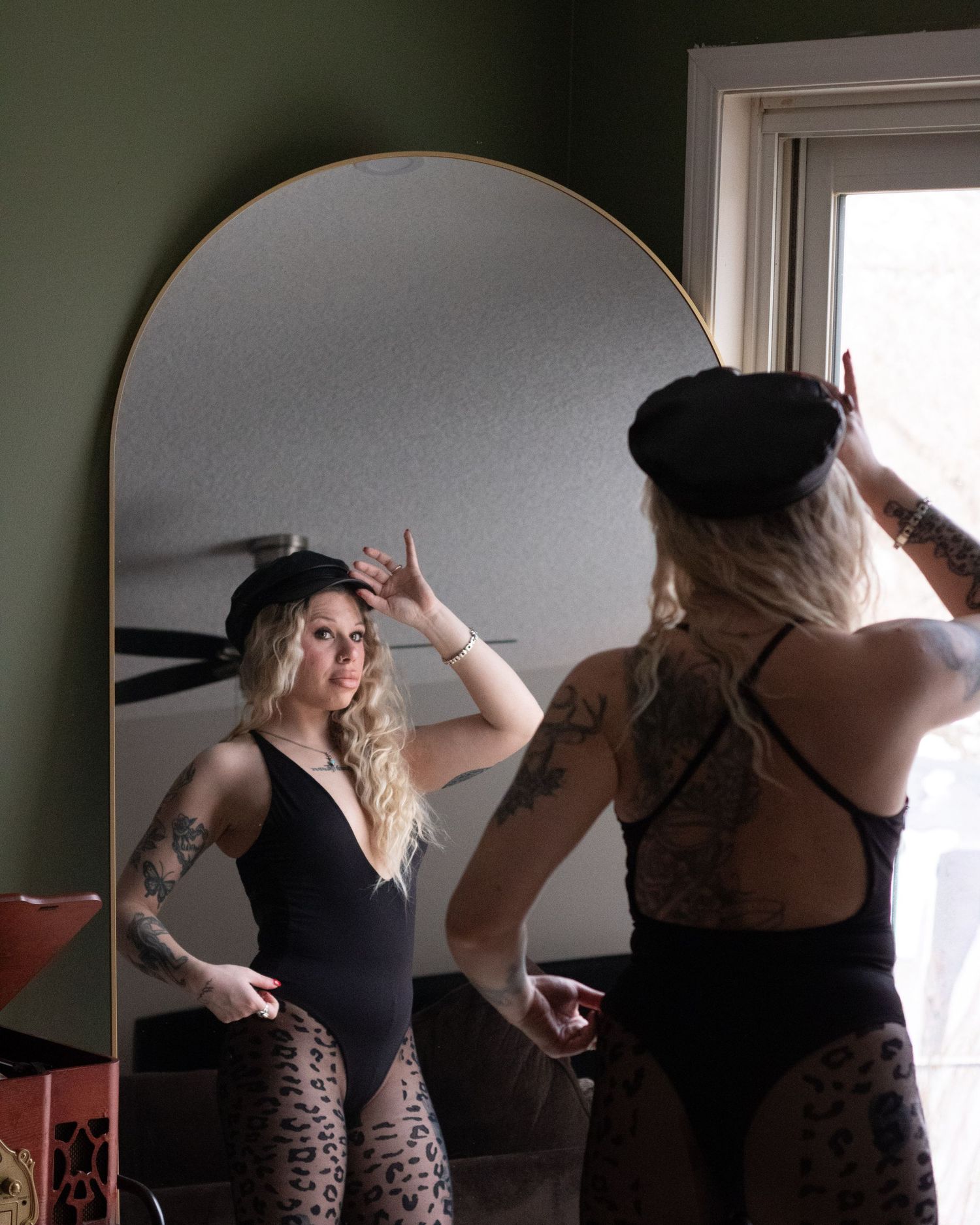 Tattooed blonde woman posing in front of an arched mirror wearing a black beret and bodysuit.