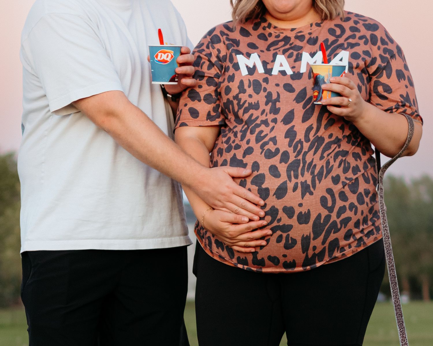 Pregnant couple holding Dairy Queen drinks outdoors, woman wearing leopard print MAMA shirt.