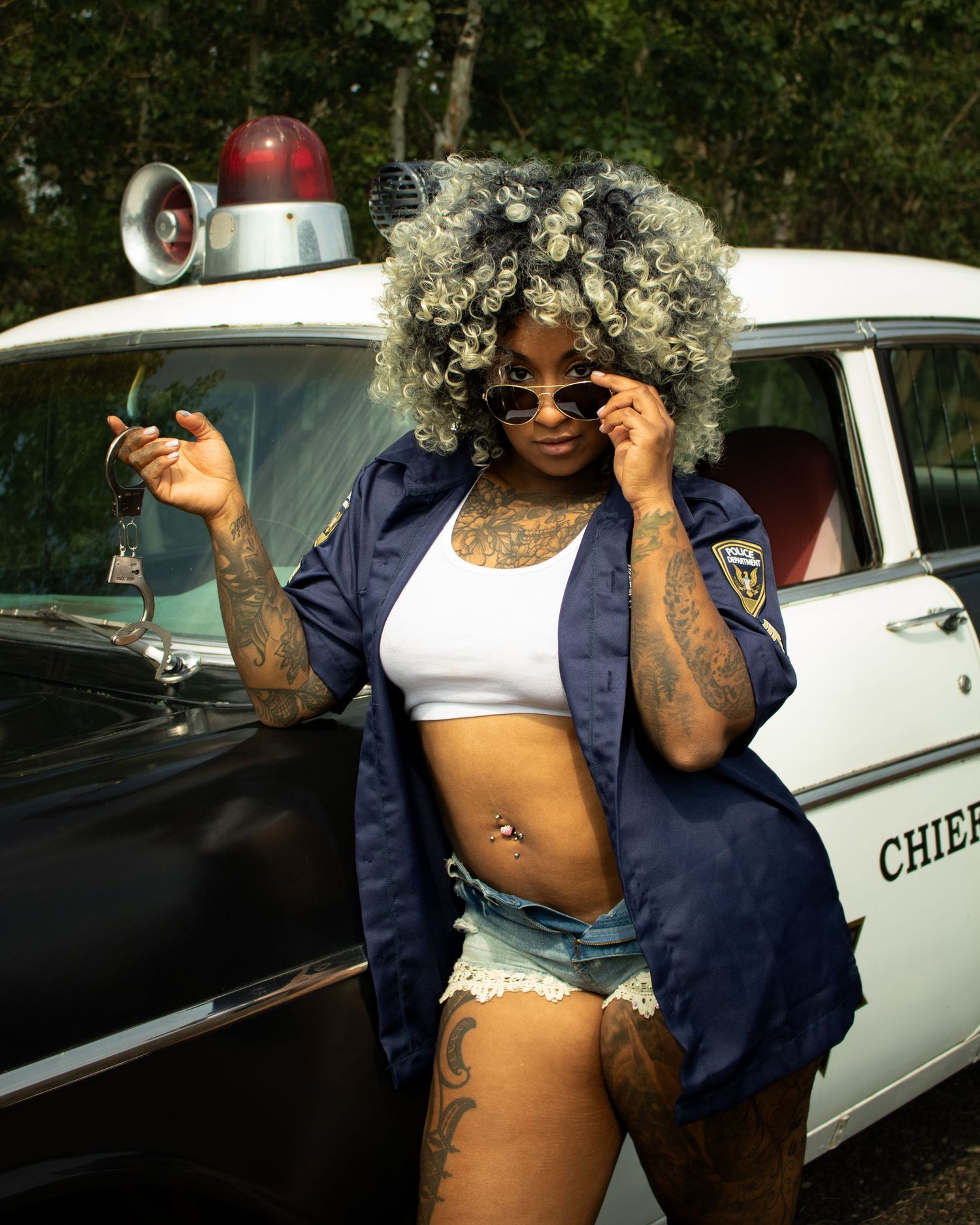A tattooed woman with curly gray hair poses in a police shirt and denim shorts beside a vintage police car.