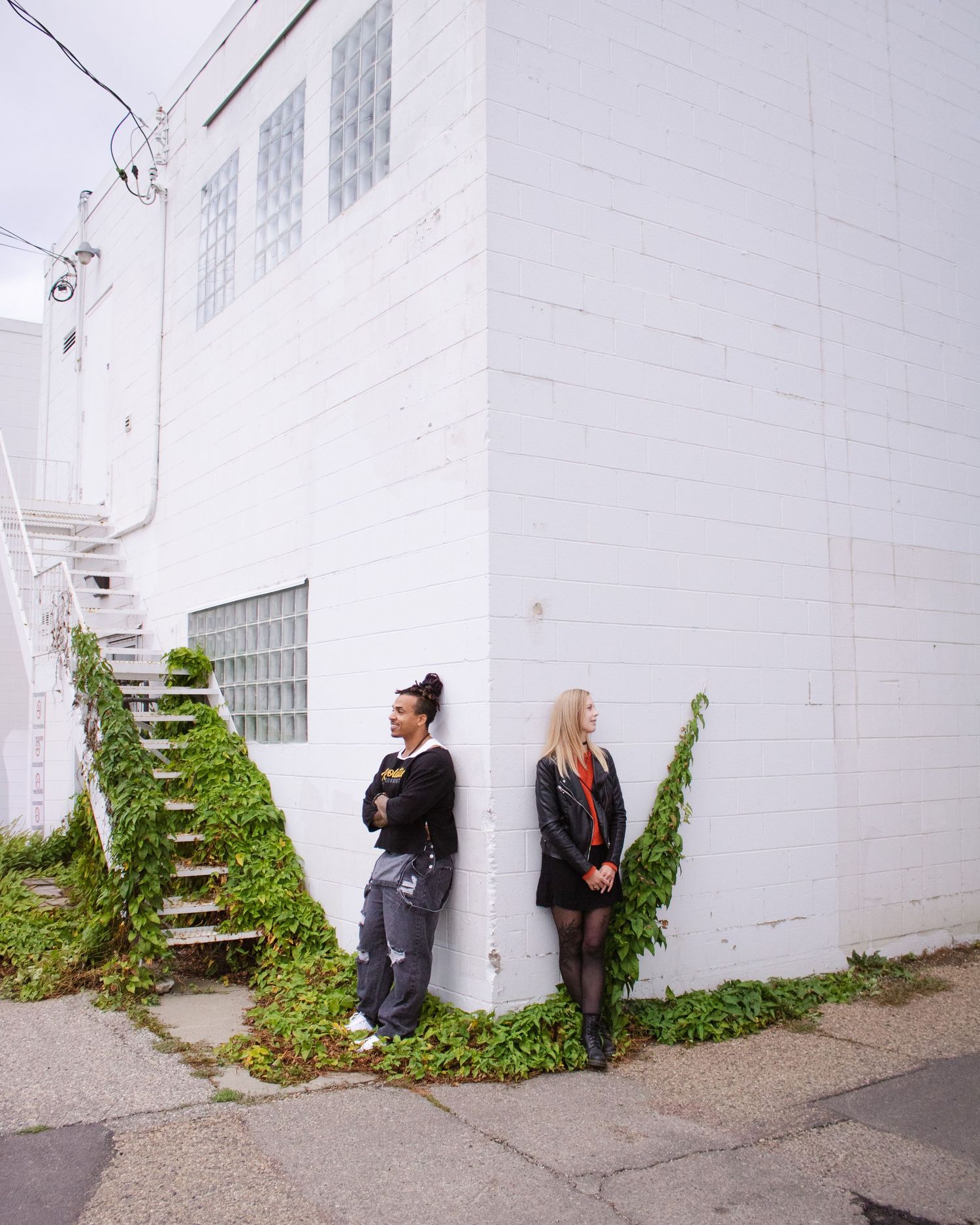 Two people standing against a white brick building with ivy, one near stairs wearing gray pants, the other in a black outfit.
