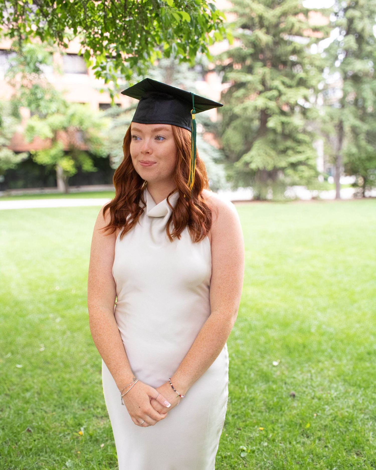 Young woman in white dress and black graduation cap smiling outdoors on green campus lawn.