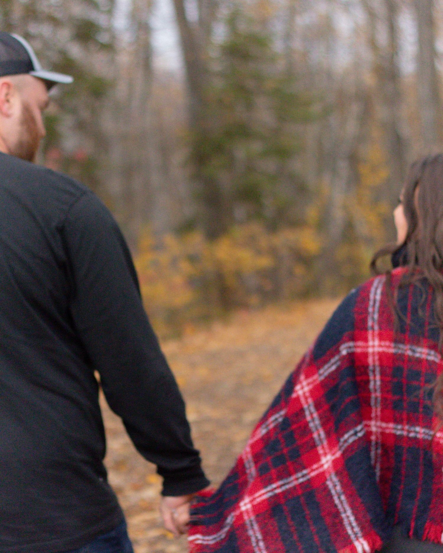 Couple holding hands walking through autumn forest path, woman wrapped in red plaid blanket among fall foliage.