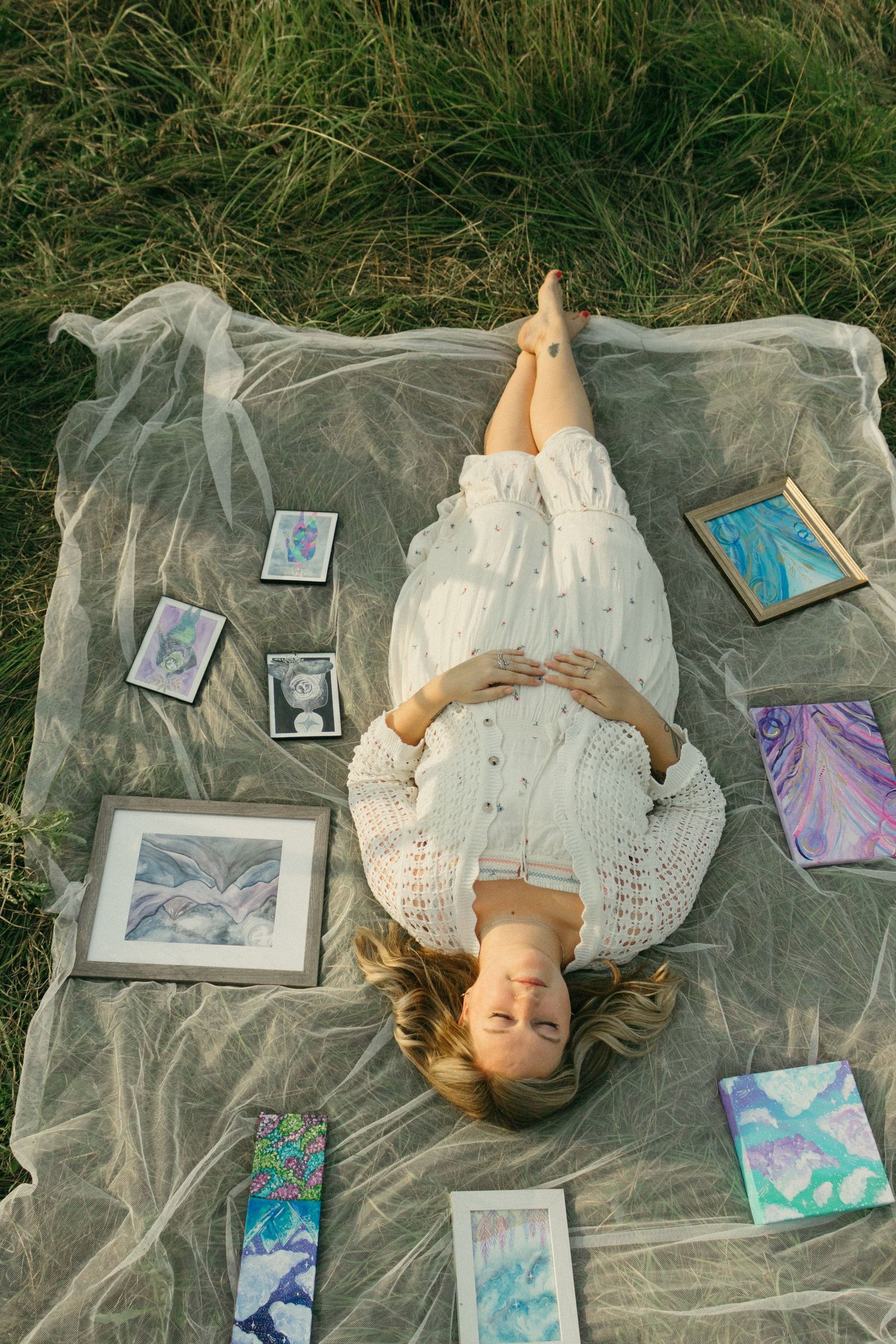Woman in white dress lying on sheer fabric surrounded by colorful artwork in a field.