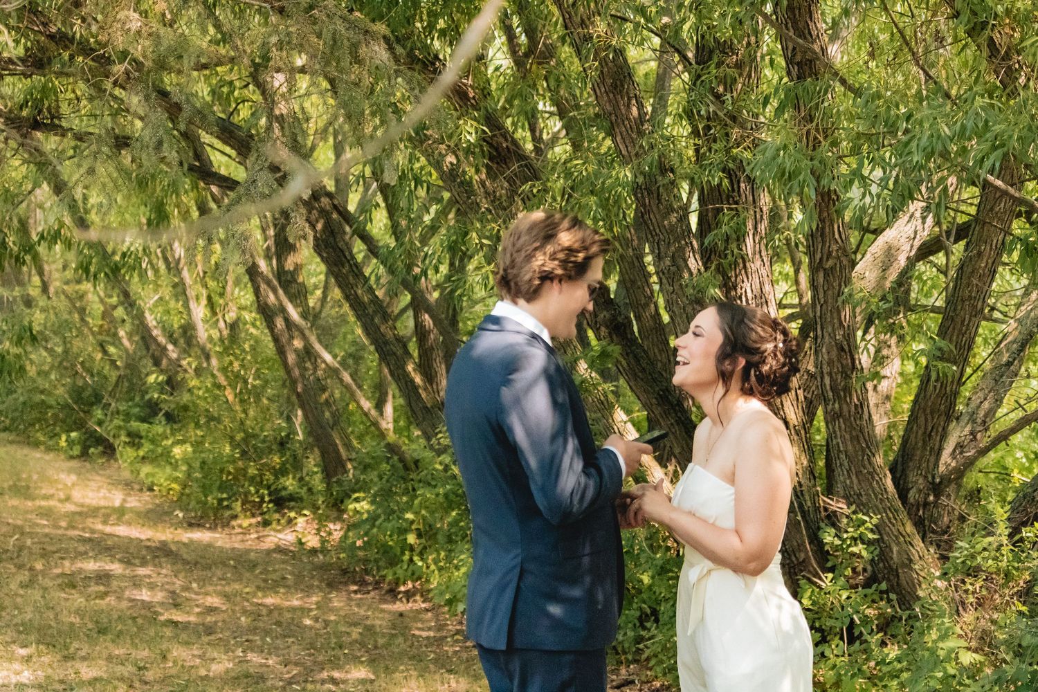 Bride and groom laughing together in a sunlit forest setting during their wedding photo session.