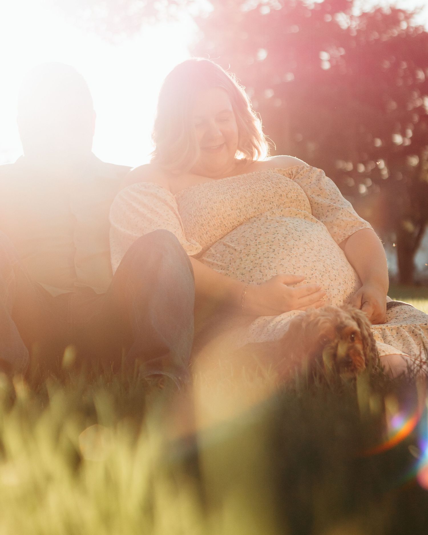 Glowing pregnant woman in lace dress sitting in sunlit grass during a dreamy maternity photo session.