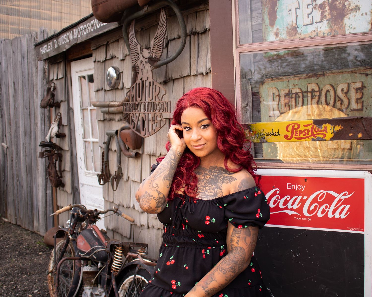 Tattooed woman with red hair in a cherry print dress posing by a vintage Coca-Cola sign.