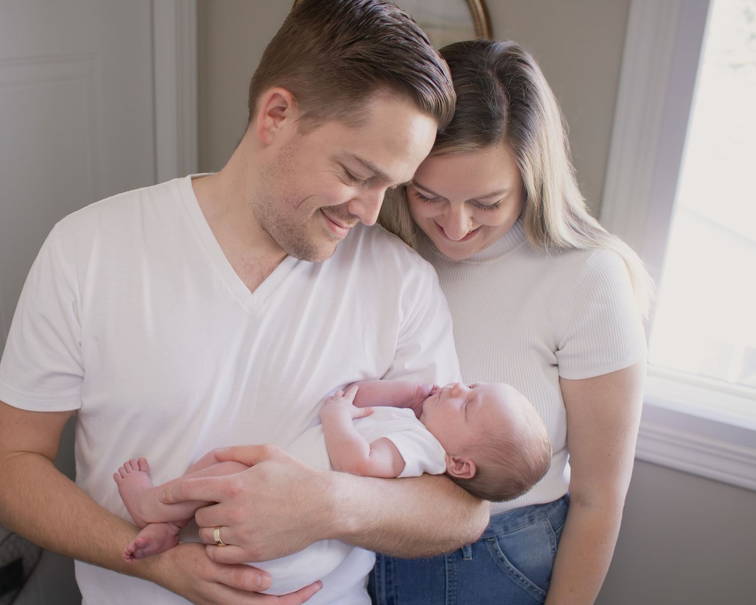 Happy couple in white t-shirts smiling at their newborn baby indoors.