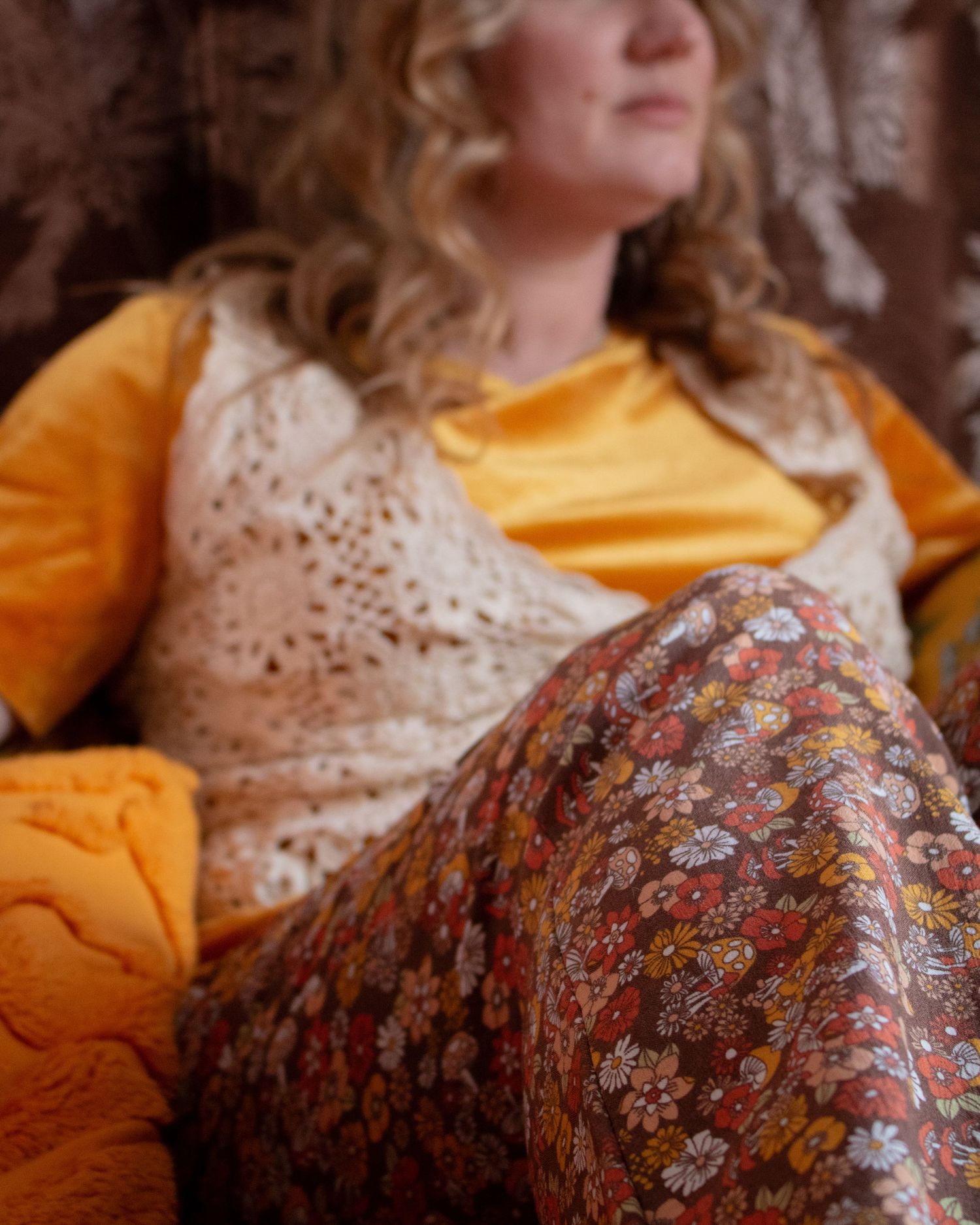 Close-up of woman in yellow top, crochet vest, and floral pants seated on an orange cushioned chair.