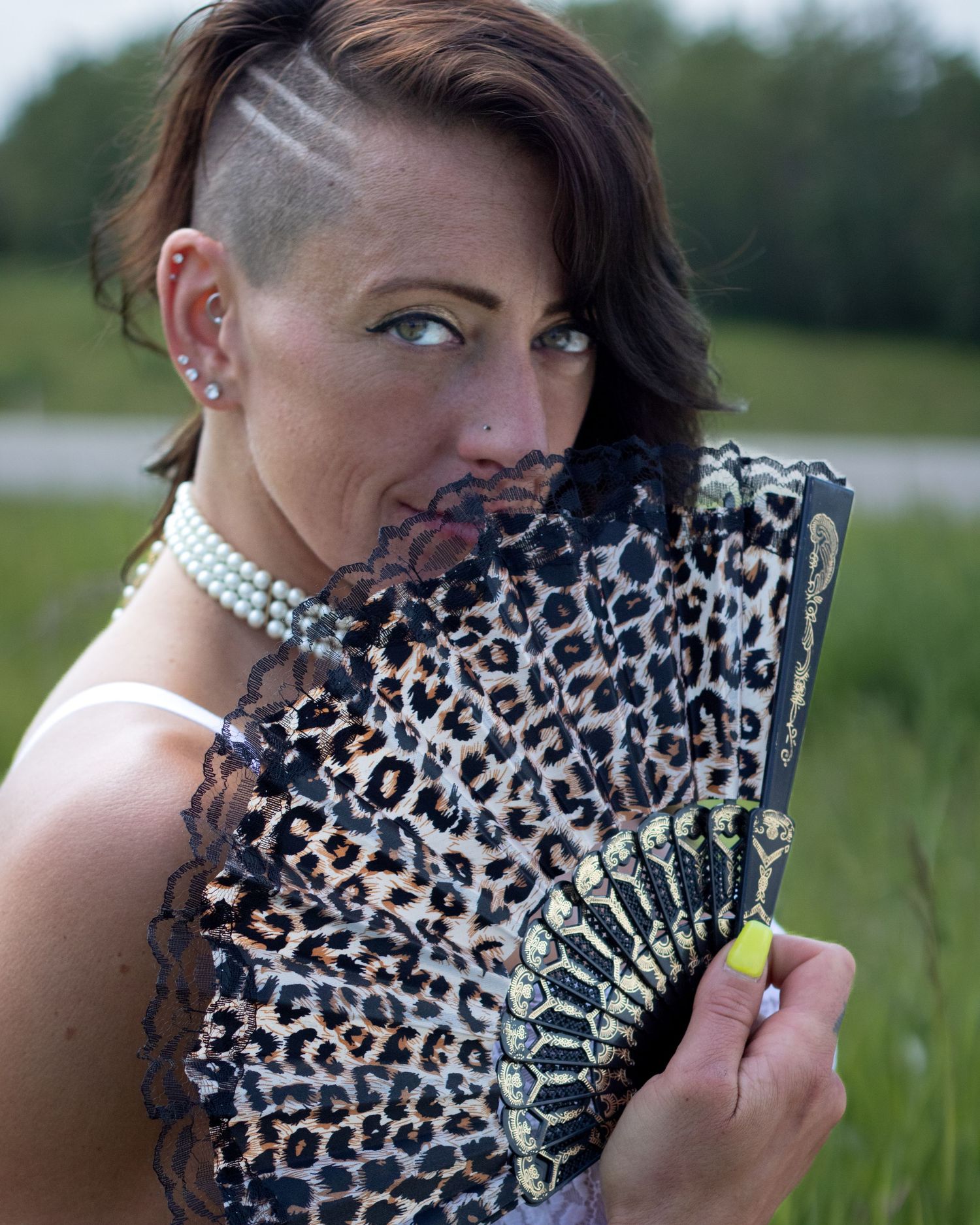 A woman with a shaved side hairstyle holds a leopard print fan near her face while wearing pearl jewelry.