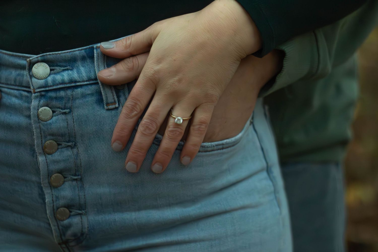 Close-up of a womans hand resting on her jeans pocket, showcasing a delicate diamond engagement ring.