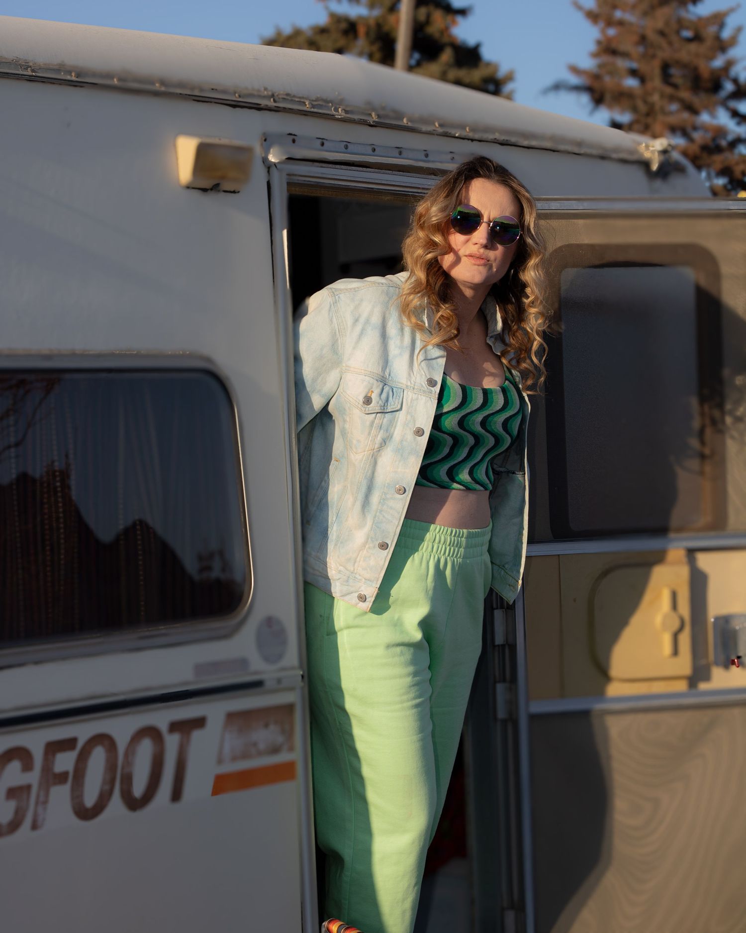 Smiling woman in mint green pants and denim jacket posing in the doorway of a Bigfoot camper.
