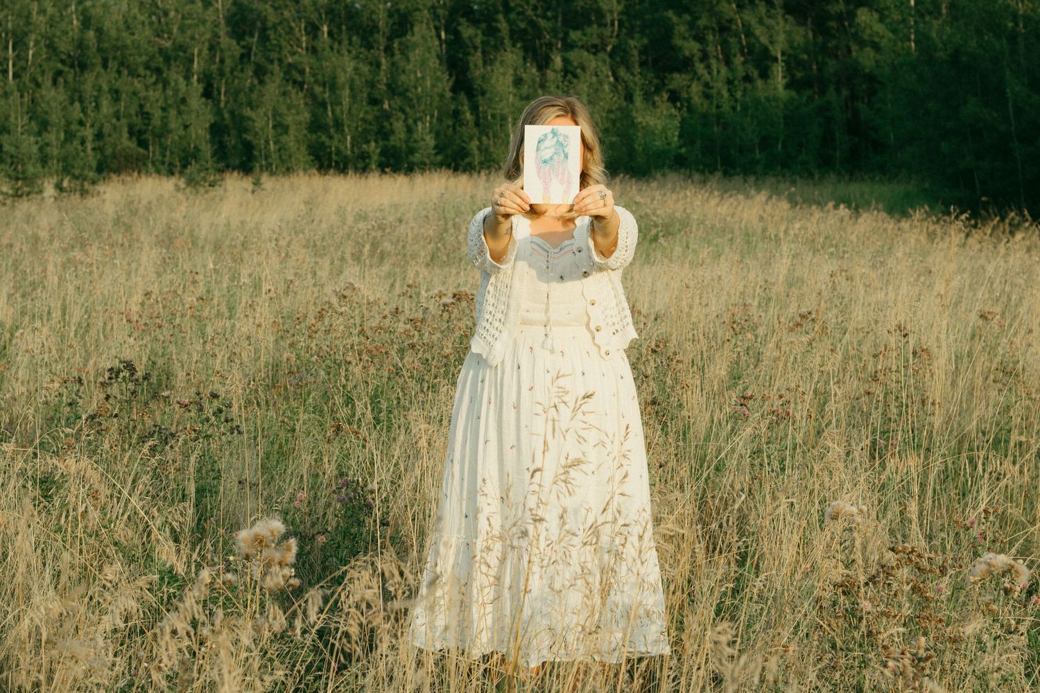 Woman in vintage white dress holding artwork over her face in a golden meadow.