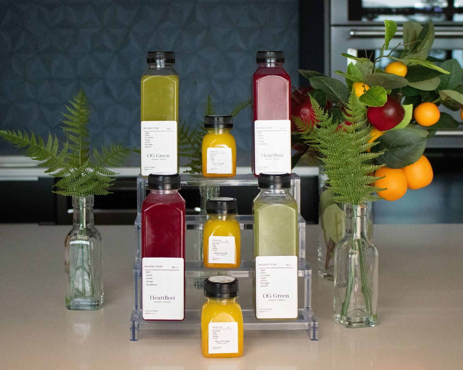 Colorful fresh cold-pressed juices displayed on tiered rack with tropical greenery and citrus decor.