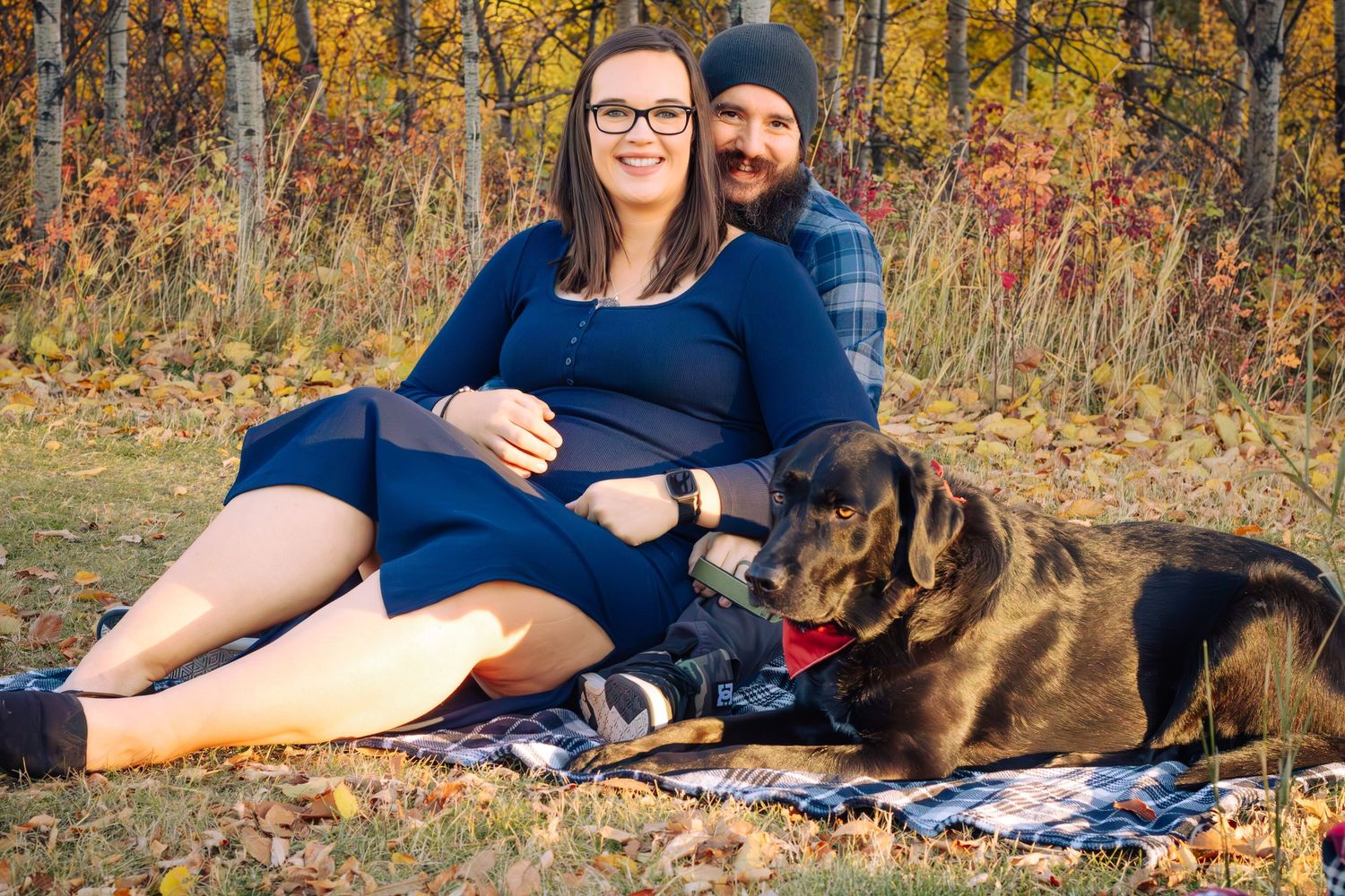Smiling pregnant couple sitting on blanket with black Labrador in autumn park setting.