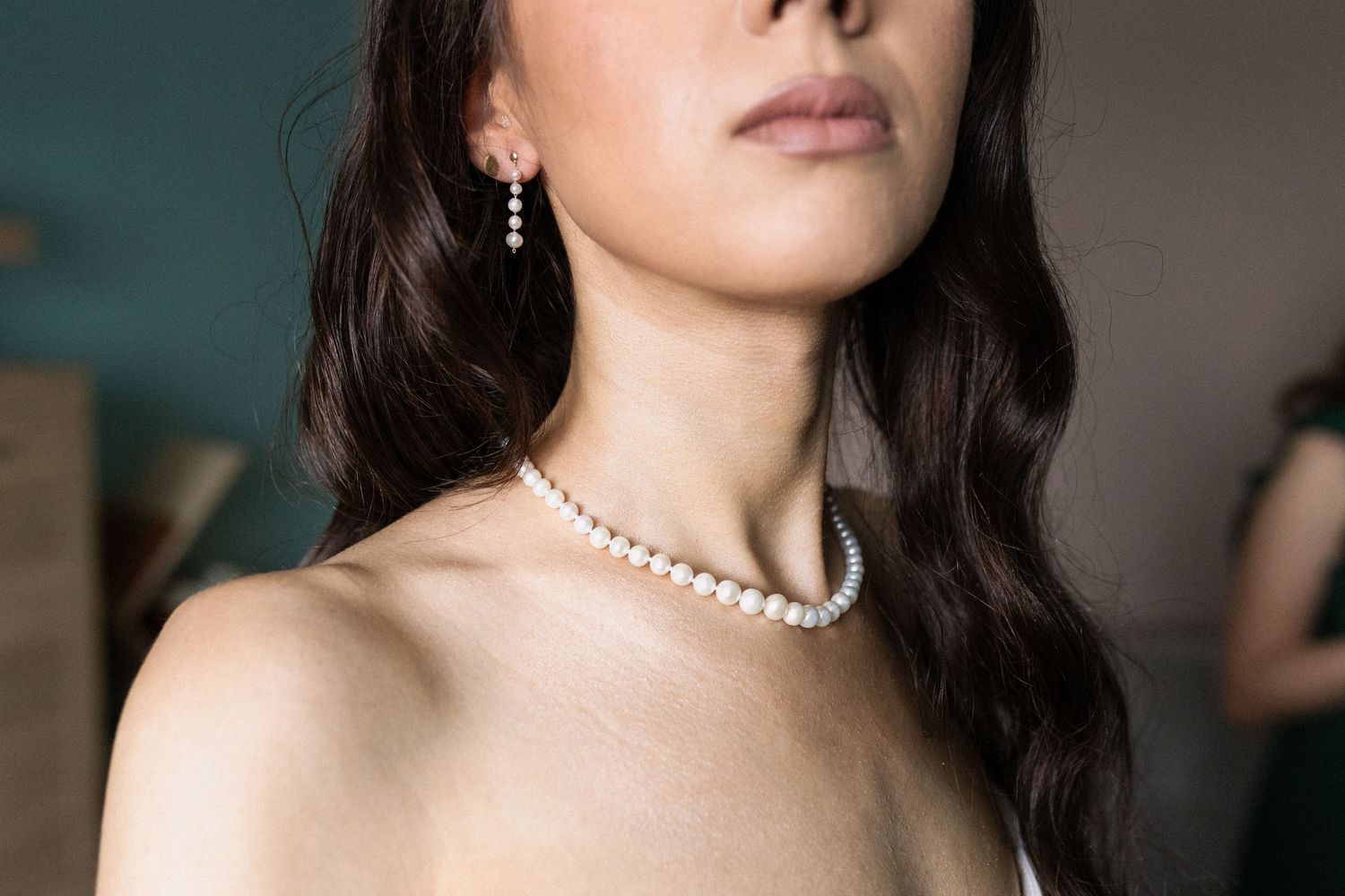 Close-up view of a pearl necklace and earrings on a person with dark wavy hair against a teal background.