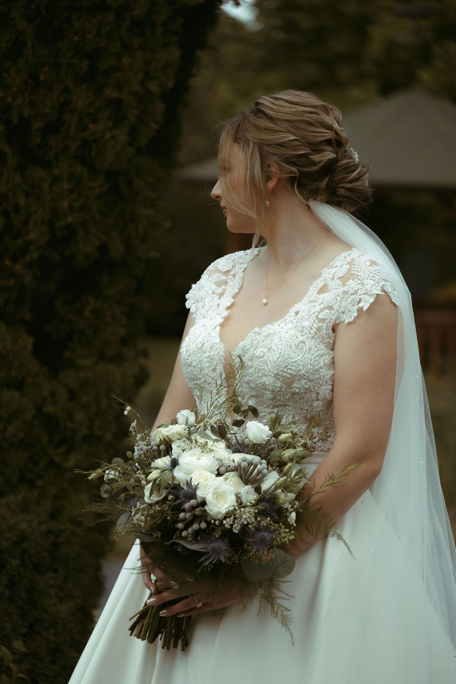 Bride wearing lace dress holds winter bouquet while looking to the side in outdoor setting.