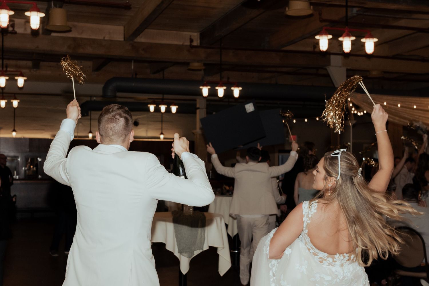 Bride and groom dancing with sparklers at wedding reception in rustic venue with string lights.