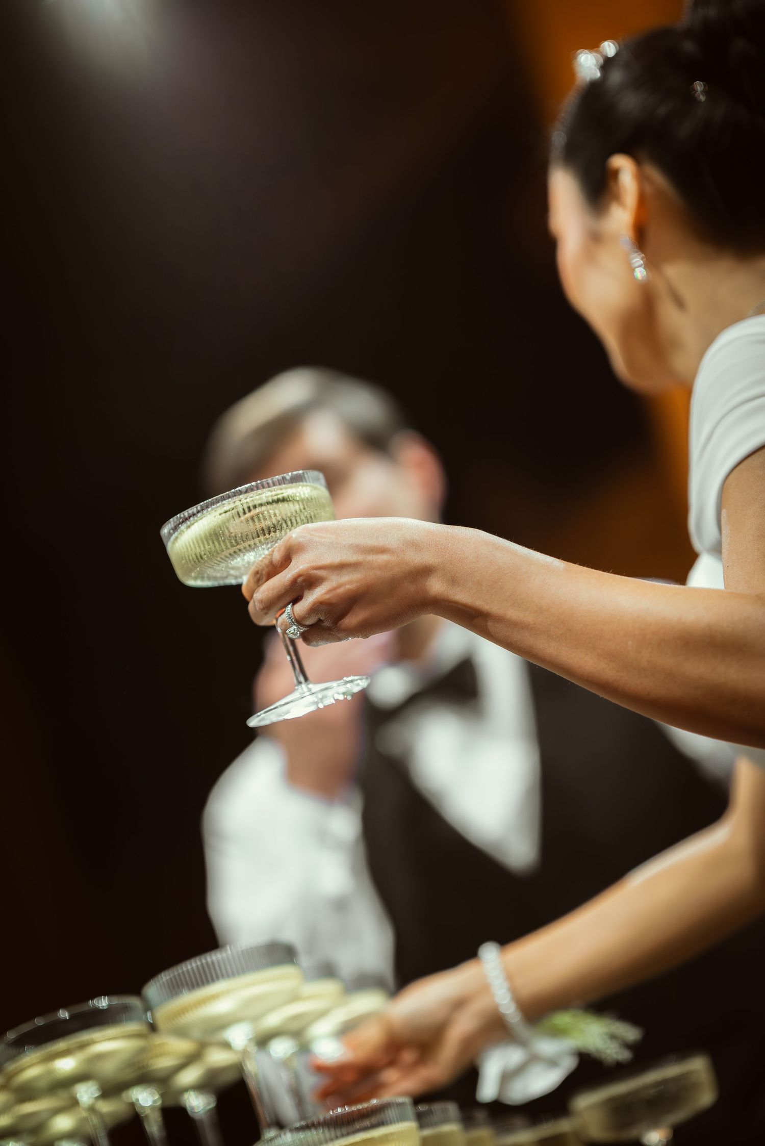 Woman in white dress holding champagne coupe glass at elegant formal event with suited staff in background.