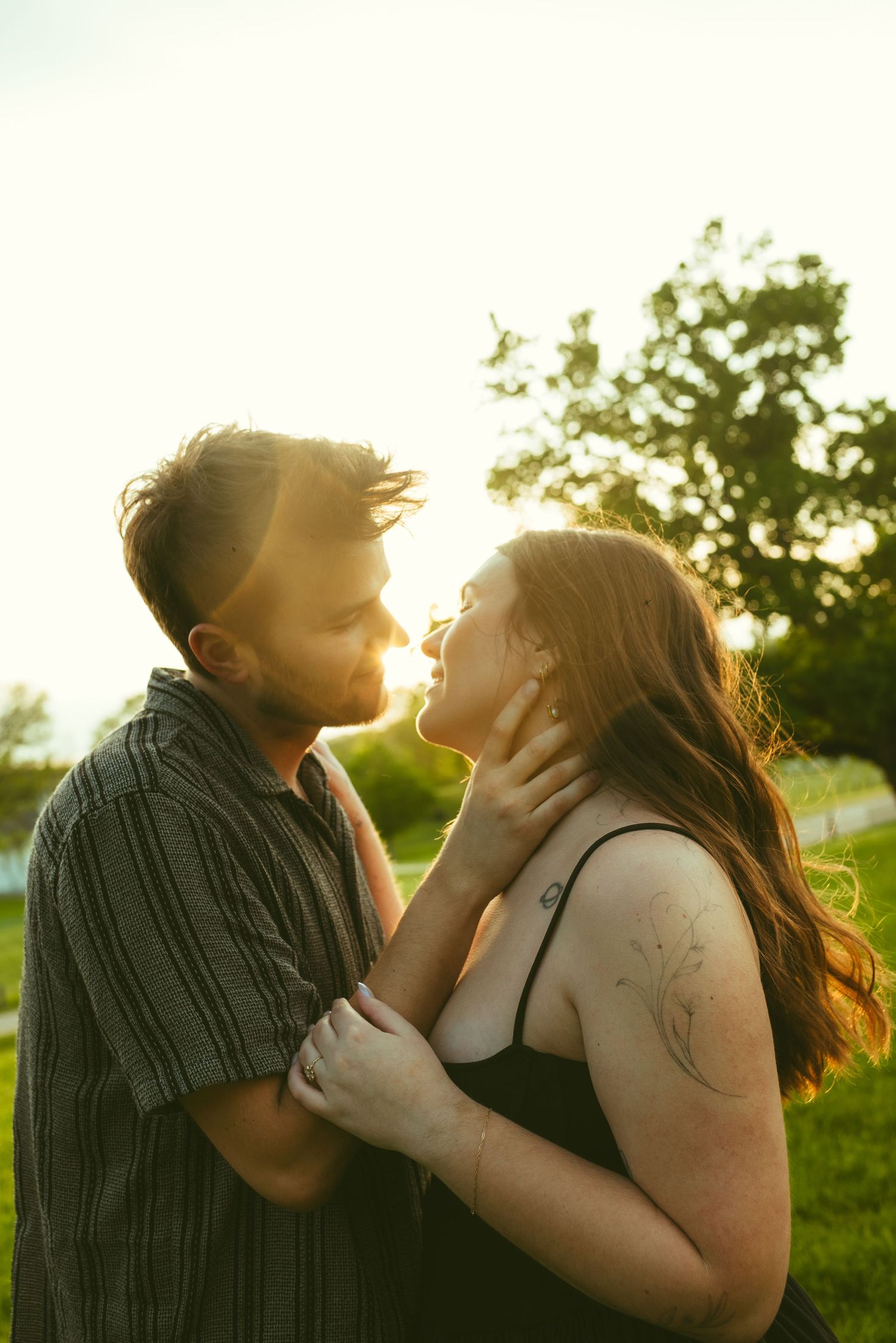 A couple shares a romantic almost-kiss outdoors at golden hour, with warm sunlight glowing behind them in a lush green park.