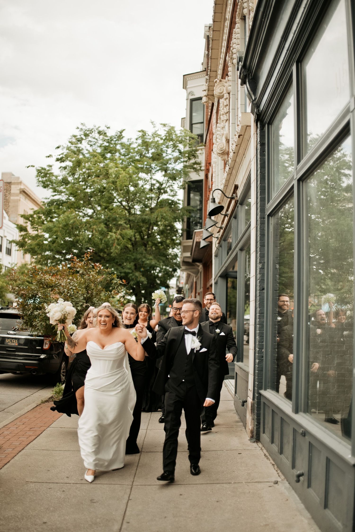 Bride and groom walk hand-in-hand with wedding party on urban sidewalk, bride holding white bouquet in elegant strapless gown.