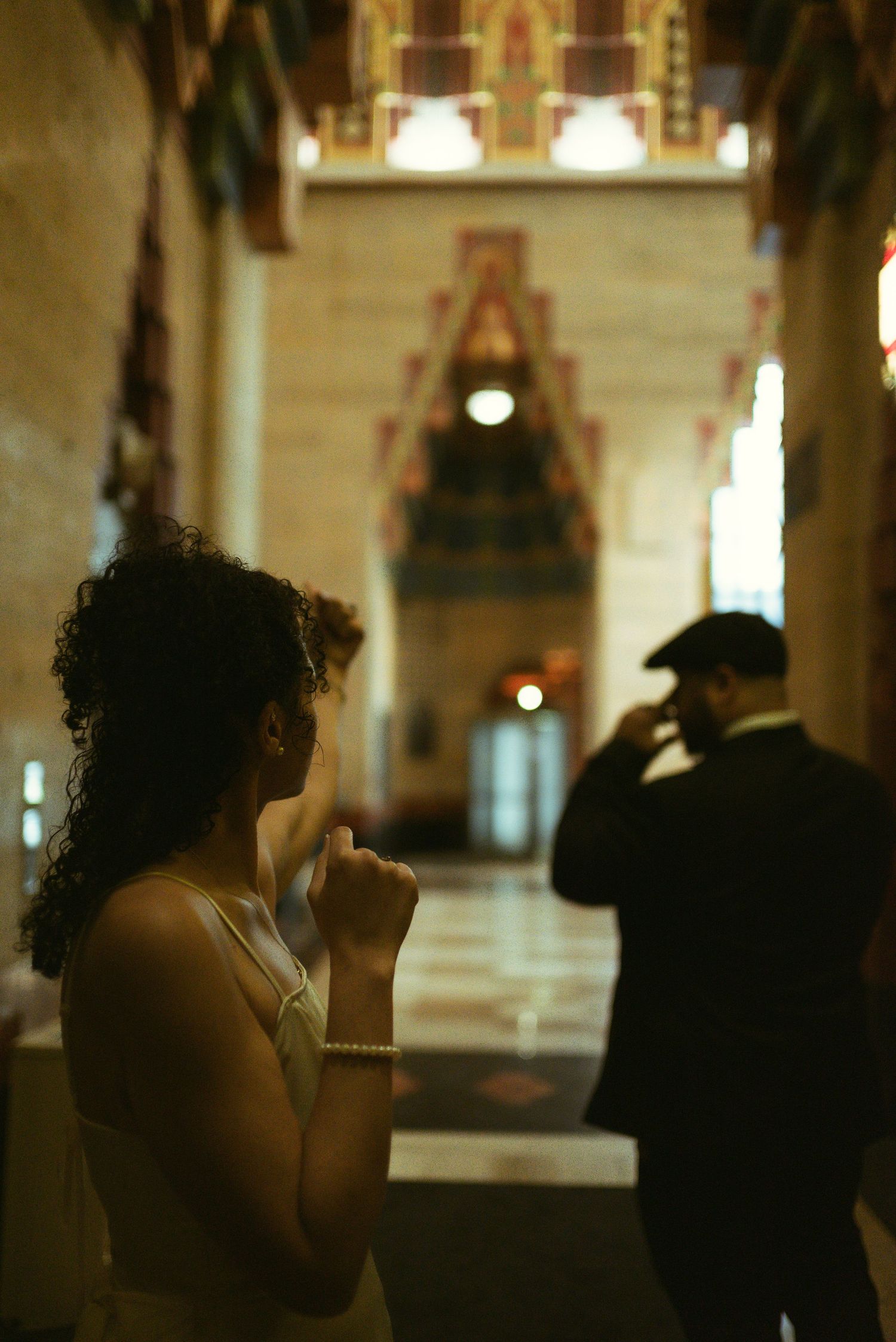 Woman in white dress praying in ornate hall with man in suit standing in background.