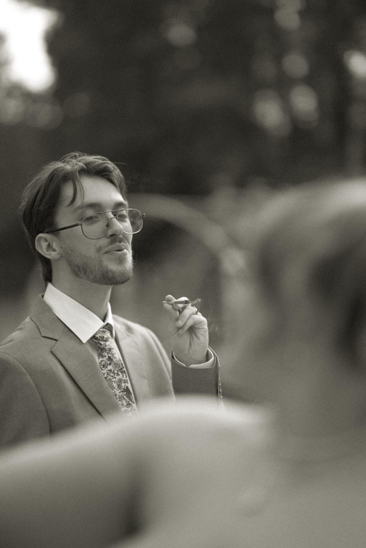 Black and white photo of a man in a suit and glasses smoking a cigar at a formal outdoor event.