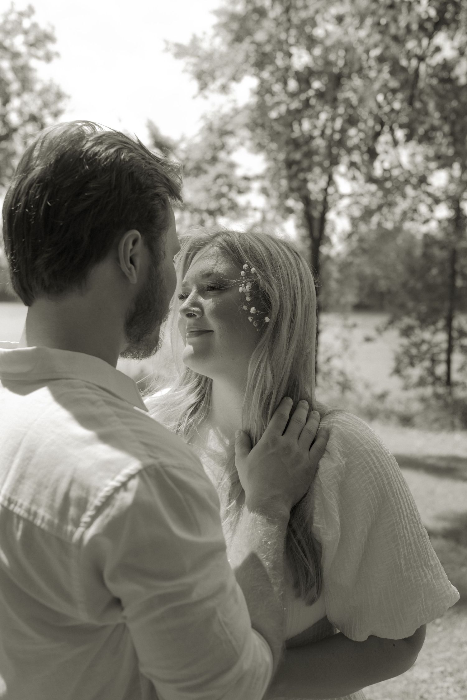 Black and white photo of a smiling blonde woman with flowers in her hair being held by a man outdoors.