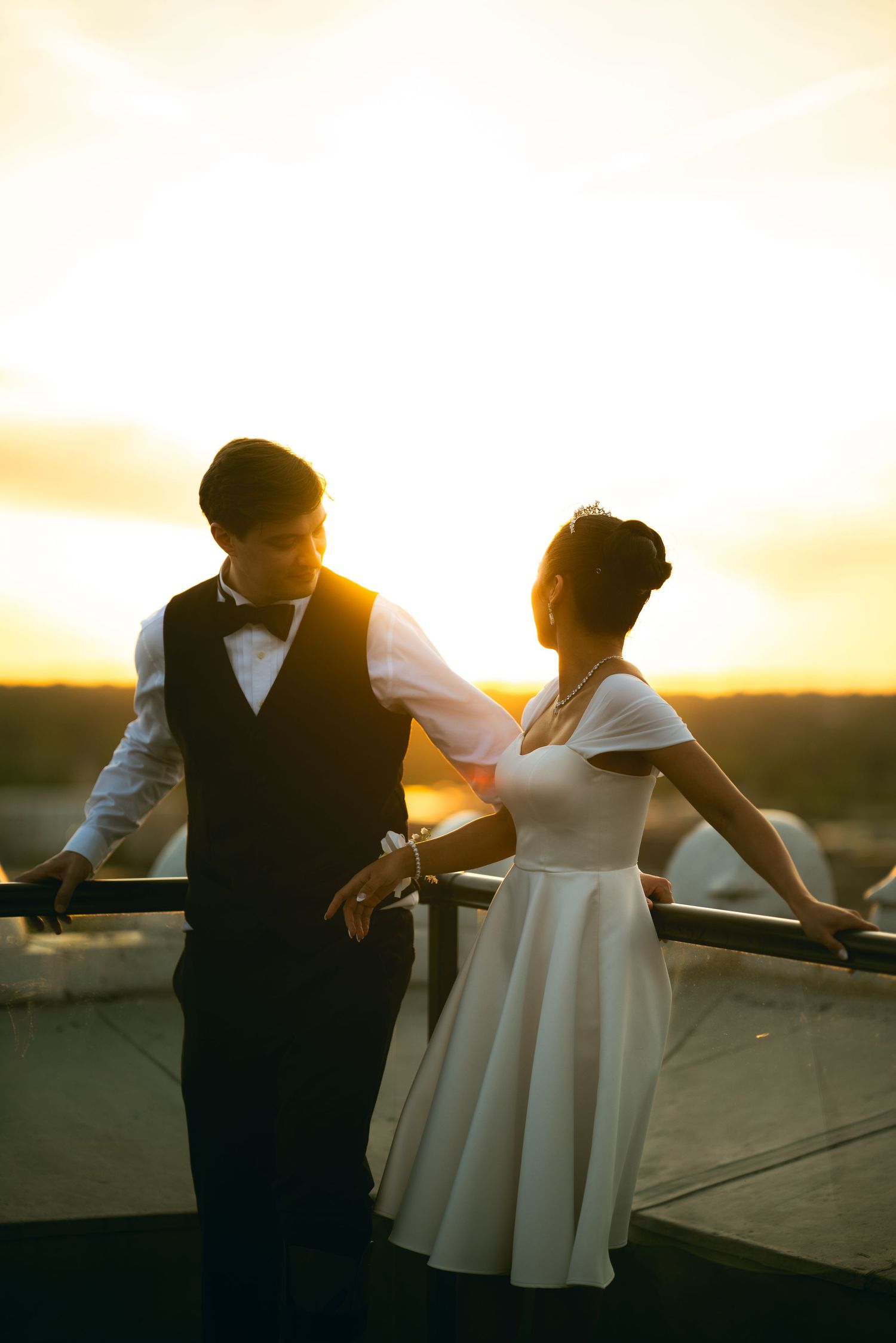 Couple in wedding attire sharing a romantic moment on a rooftop at golden sunset, man in vest and woman in white dress.