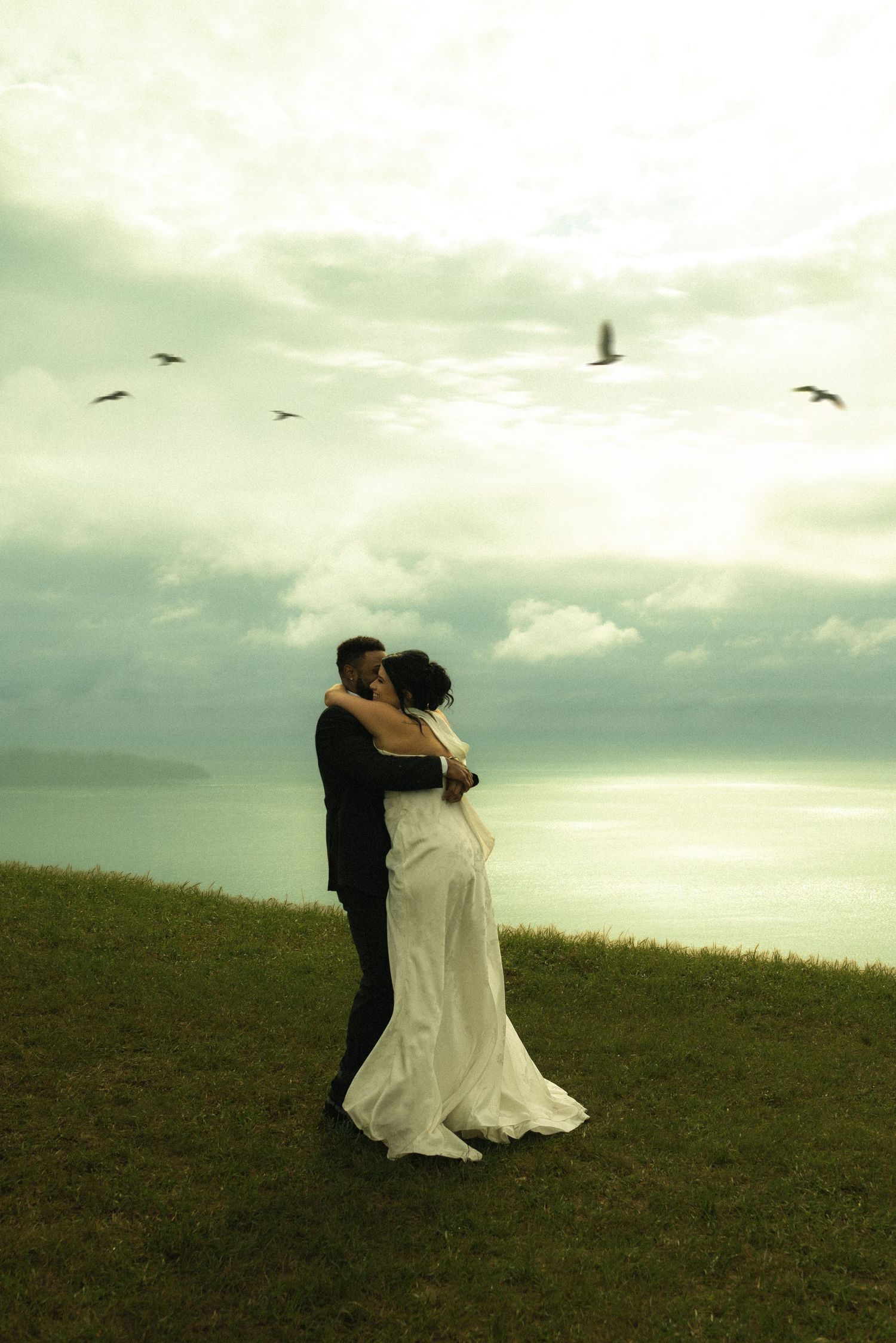 A bride and groom embrace on a grassy cliff overlooking the ocean under a dramatic cloudy sky with birds flying above.