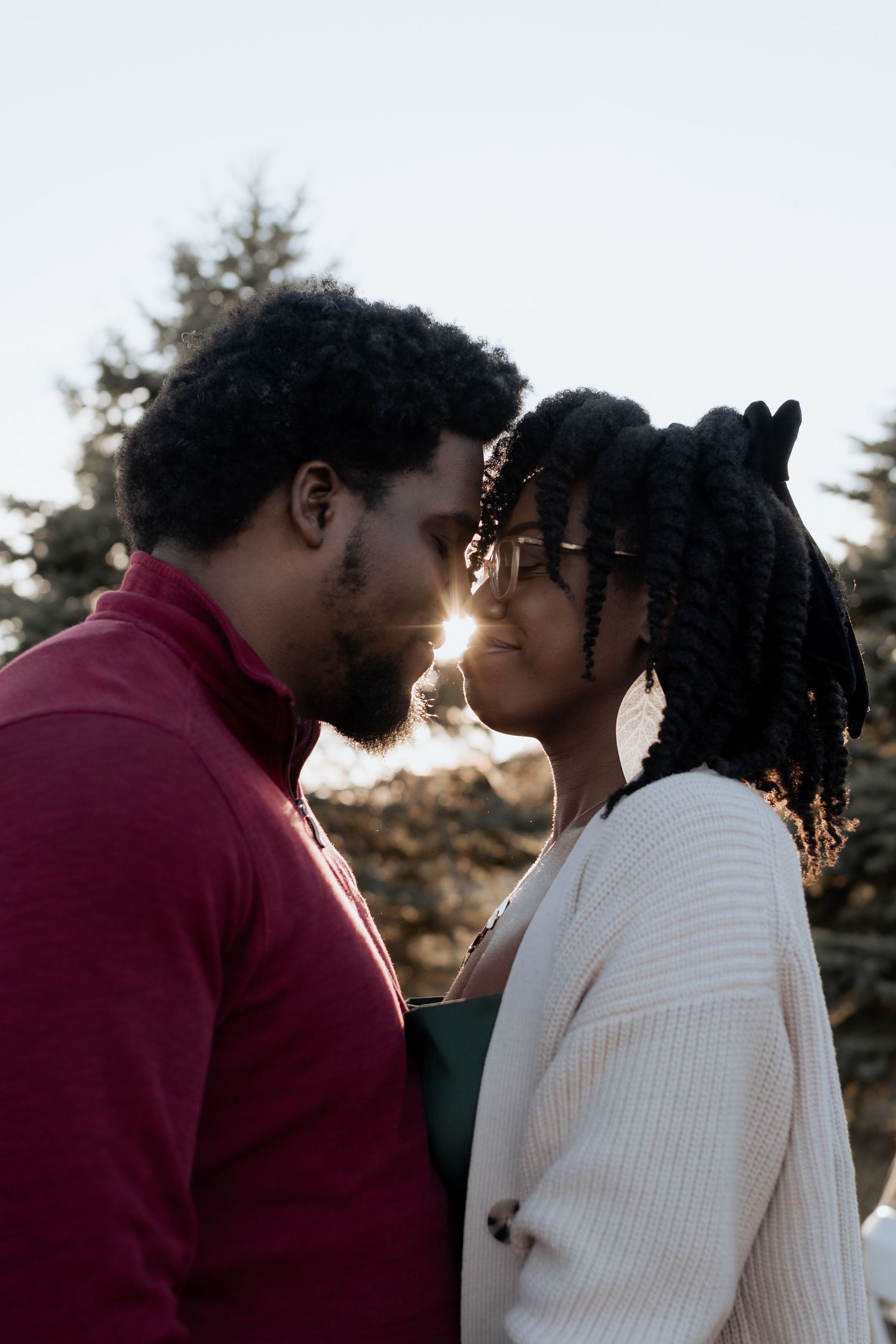 A couple sharing an intimate moment outdoors, with sunlight glowing between them, surrounded by trees.