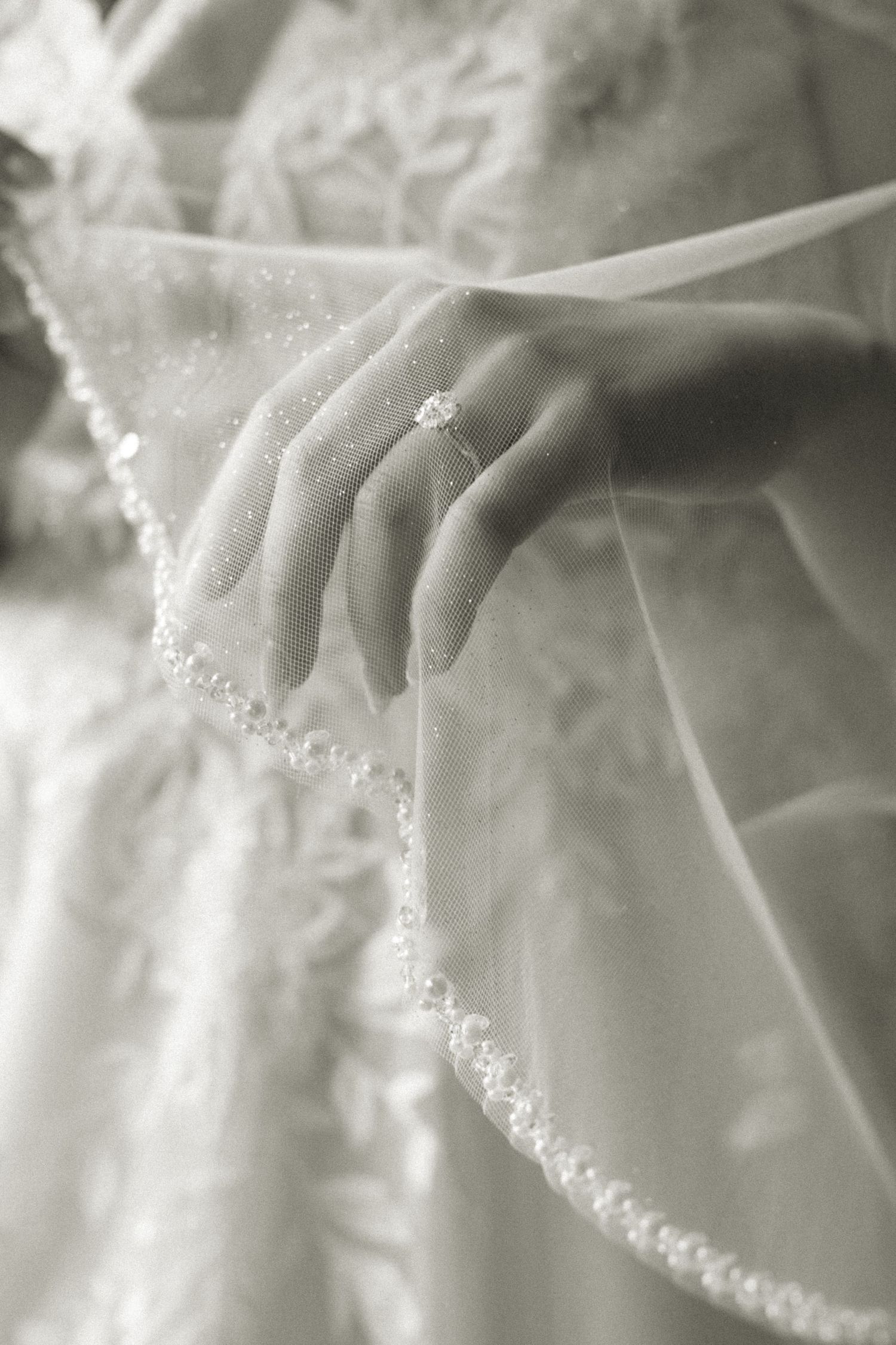 A delicate wedding veil with beaded trim frames an engagement ring on a hand against soft white fabric.